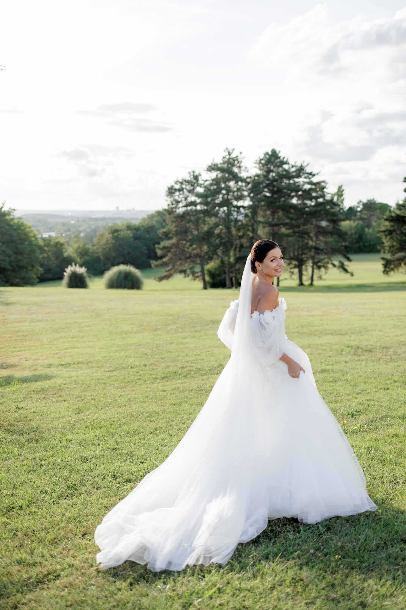 A bridal portrait taken outdoors on the grounds of what appears to be a large estate or chateau, with an expansive lawn and a wide panoramic view in the background. The bride, a dark-haired woman with her hair pinned up, is glancing back over her shoulder toward the camera with a smile. She wears a white voluminous ball gown with an off-the-shoulder neckline, puff sleeves, and a long tulle train spread across the grass, paired with a cathedral-length veil. The composition is a full-length portrait shot at a slight distance, capturing the full spread of the dress and train against the open lawn setting.