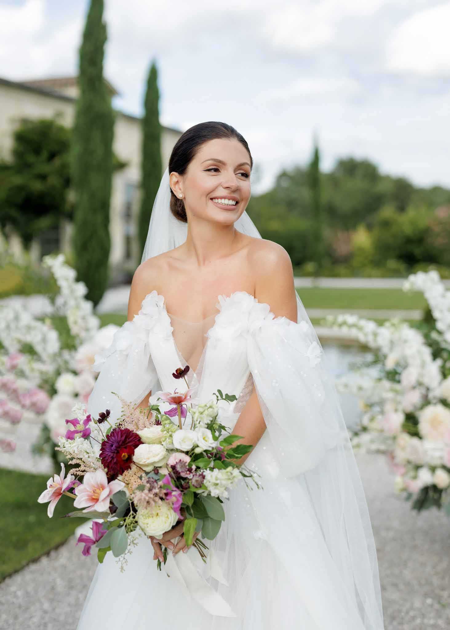 A bridal portrait taken outdoors in the formal garden of what appears to be a French villa or chateau, with tall cypress trees and a reflecting pool visible in the background. The bride wears an off-the-shoulder white gown with three-dimensional floral appliqué detailing on the sleeves and a deep V-neckline, paired with a shoulder-length cathedral veil and pearl stud earrings, her dark hair pulled back in a low updo. She holds a large, loosely arranged bouquet featuring burgundy dahlias, blush and hot pink lilies, cream garden roses, white ranunculus, dark chocolate cosmos, astilbe, and eucalyptus foliage. Behind her, large white floral arch installations frame the path, and the overall styling aesthetic is classic with a romantic, garden-influenced approach to florals.