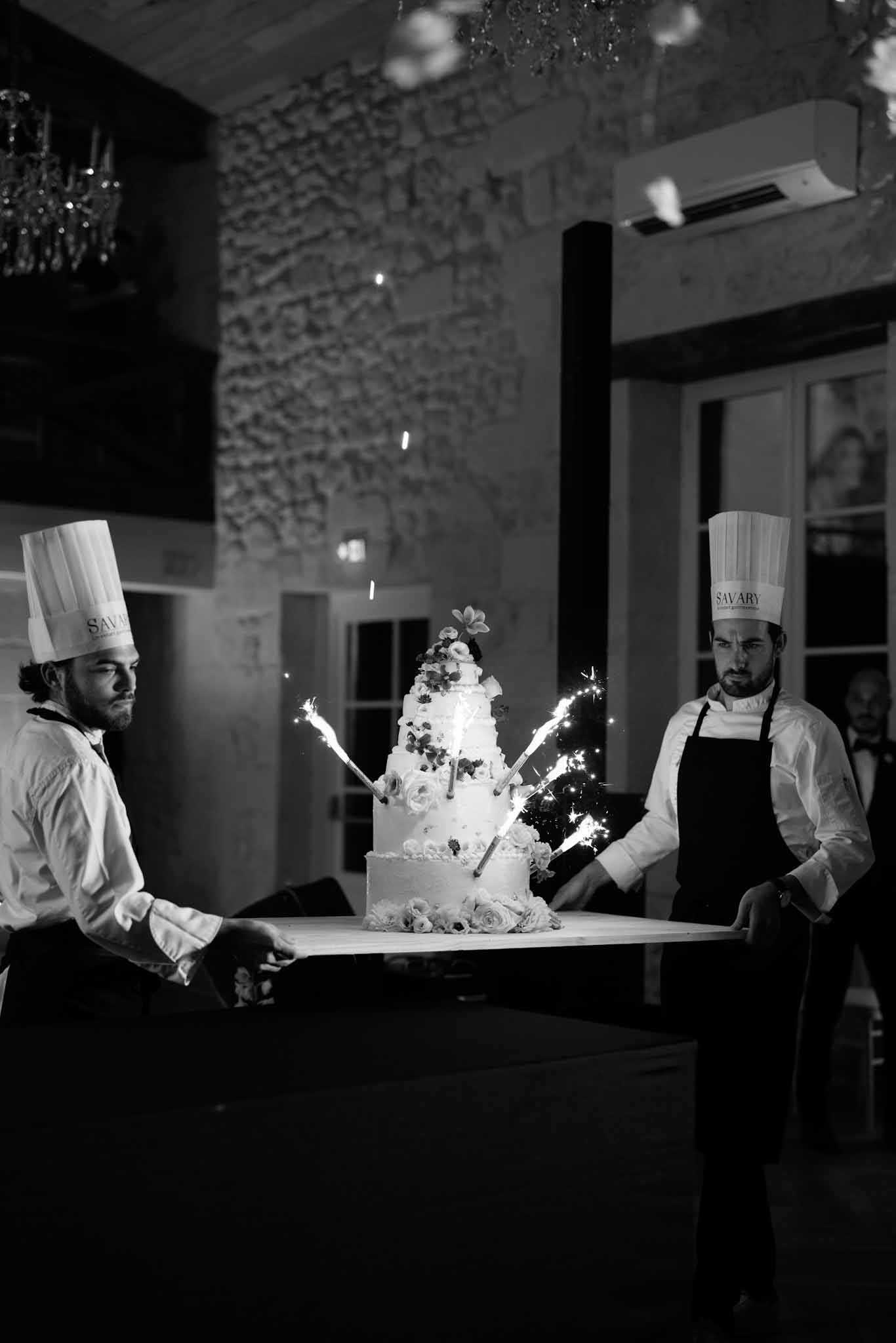 This black-and-white image captures the wedding cake presentation moment during an indoor evening reception. Two male pastry chefs from the catering company Savary, wearing tall white toques and dark aprons over white shirts, carry a large multi-tiered wedding cake on a white board toward the guests. The cake, which appears to be approximately four tiers, is decorated with floral clusters at the base and top and has multiple lit sparklers shooting bright sparks in multiple directions. The setting is an indoor venue with exposed stone walls and a chandelier visible overhead, giving it a classic French château feel. The image is shot in medium wide portrait format, with high contrast black-and-white tones that emphasize the bright sparkler light against the dark reception room.