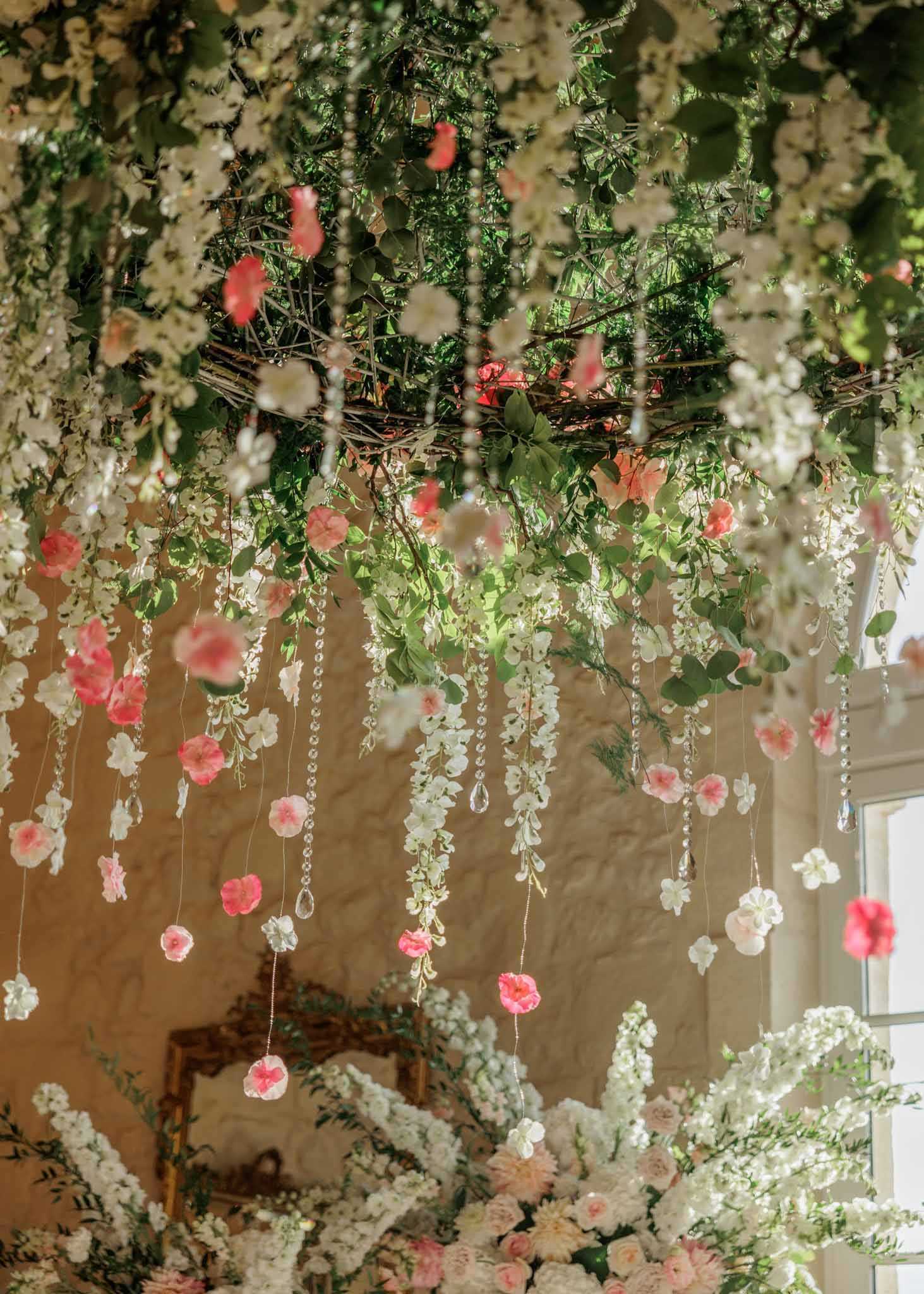 Close-up detail shot of an overhead floral installation inside a stone-walled venue room. The hanging structure is built on a twig-and-branch frame densely packed with green foliage, white wisteria-style trailing flowers, and coral-pink blooms, with crystal bead strands and teardrop pendants cascading downward. Individual coral and blush pink flowers appear to float along the crystal strands at varying heights. In the lower portion of the frame, large ground-level floral arrangements of white delphinium, blush pink roses, and green foliage are partially visible. The decor palette is white, blush, and coral pink against a warm stone wall, with natural daylight entering from a window to the right, creating a romantic garden-inspired indoor aesthetic.