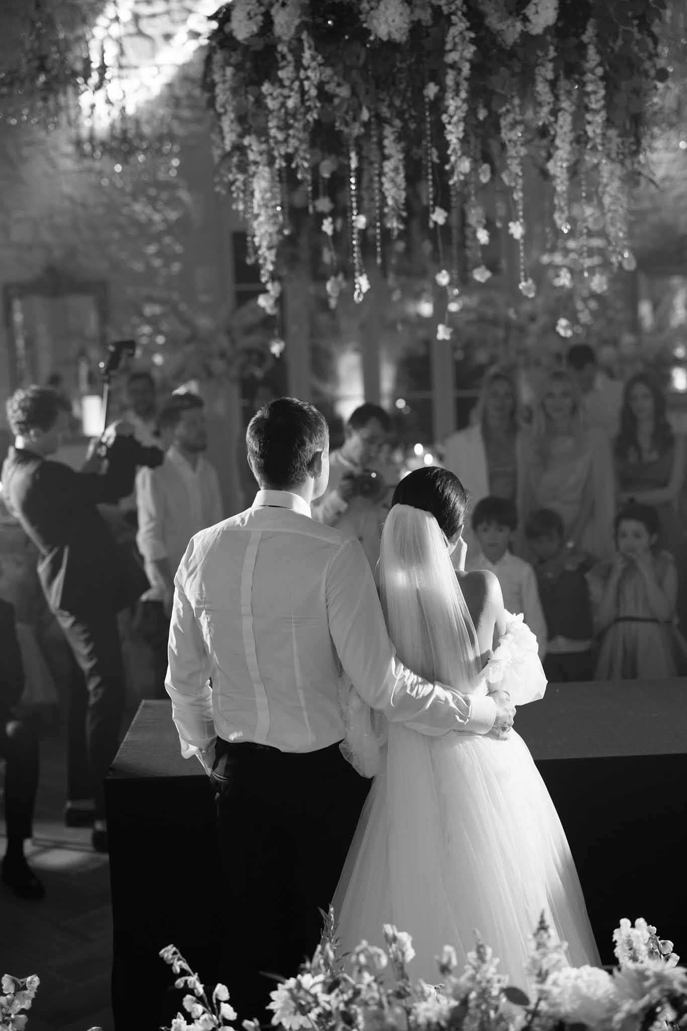 Black-and-white portrait-style shot of a bride and groom standing at the altar during an indoor wedding ceremony, photographed from behind. The groom wears a white dress shirt and dark trousers, while the bride wears a full tulle gown with a long veil and holds a bouquet. The ceremony space features an elaborate overhead installation of cascading florals and hanging crystal or bead strands, creating a dense canopy above the couple. Floral arrangements are also visible in the foreground at the base of the altar. An officiant stands facing the couple in the middle ground, with approximately 15–20 seated and standing guests visible on both sides in the background. The interior space appears to have stone walls with warm uplighting visible in the mid-tones, suggesting a barn or chapel venue. The image has soft, warm contrast with bright highlights on the bride's veil and gown against the darker background.