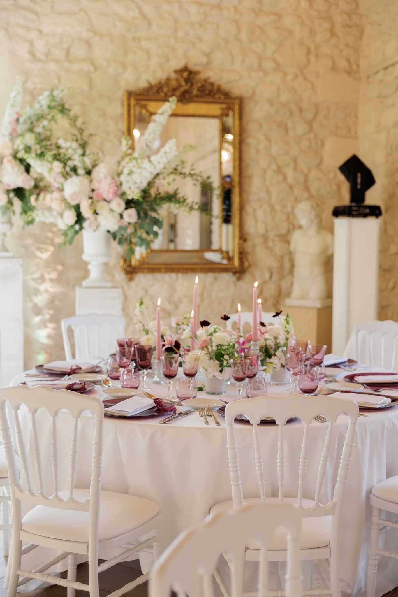 A detail shot of a wedding reception tablescape set inside an ornate indoor venue with textured cream stone walls. The round table is dressed with a white linen tablecloth, deep burgundy charger plates, gold flatware, and mauve-pink glassware including both wine glasses and smaller tinted glasses. The centerpiece features a low arrangement of white ranunculus, blush garden roses, dark burgundy anemones, and greenery, flanked by lit dusty pink taper candles in individual holders. In the background, a tall white urn holds a voluminous floral arrangement of blush peonies, white delphiniums, and trailing greenery, positioned near a gold ornate mirror mounted on the wall; a white pedestal with a classical bust is visible to the right. White French Napoleon-style chairs surround the table, contributing to a classic, romantic French interior aesthetic.