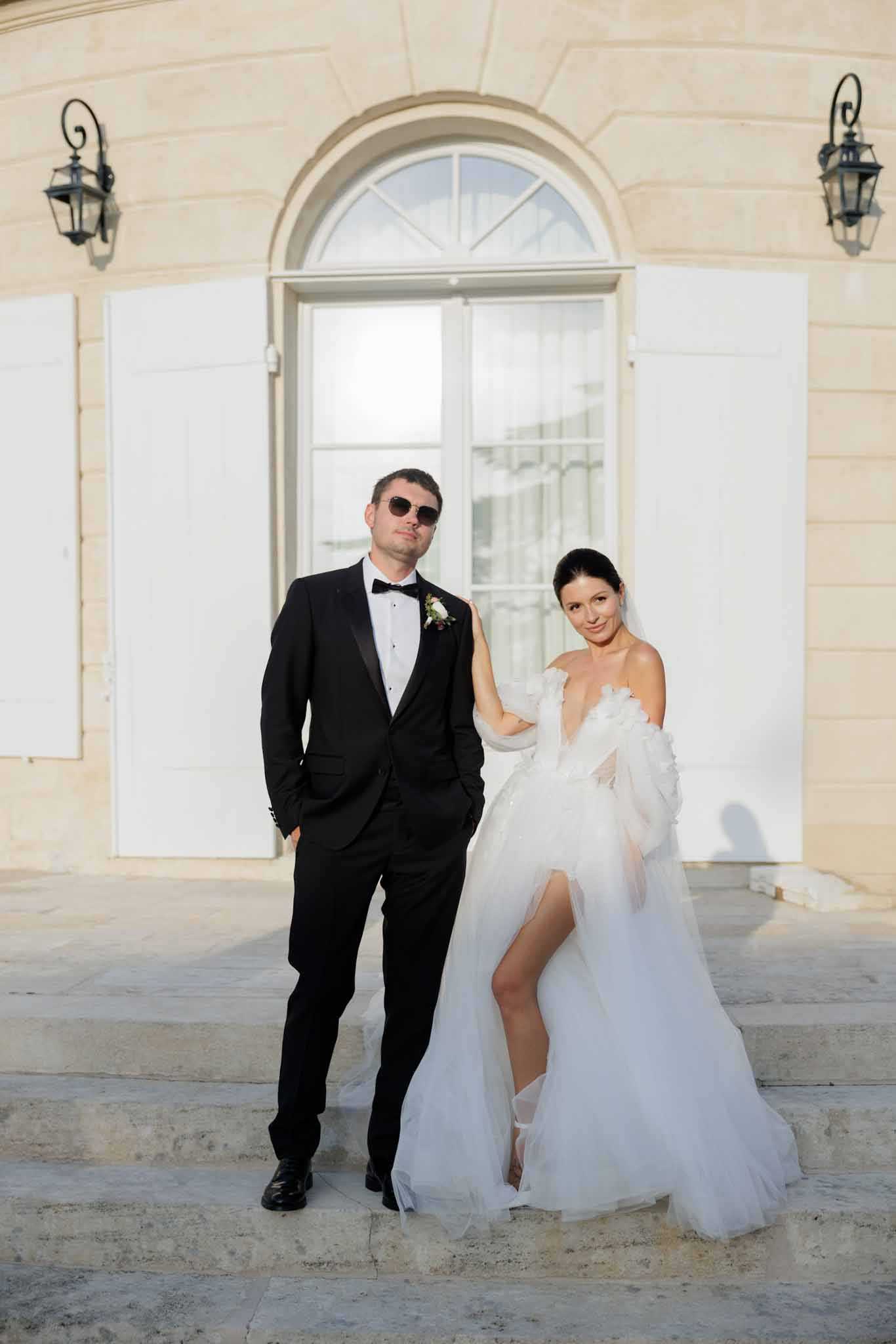 A couple portrait taken outdoors on the stone steps of a French chateau, with the limestone facade, arched window, white shutters, and wrought-iron wall lanterns visible behind them. The groom wears a black tuxedo with a bow tie and dark sunglasses, and has a small white and burgundy boutonniere on his lapel; he stands with one hand in his pocket while the bride rests her hand on his arm. The bride wears a white off-shoulder tulle ballgown with a deep V-neckline, 3D floral appliqué detailing at the shoulder, an off-shoulder tulle wrap, and a high front slit. The styling is modern and fashion-forward, combining classic black-tie formalwear with an editorial edge. This is a full-length couple portrait shot in warm late-afternoon light.