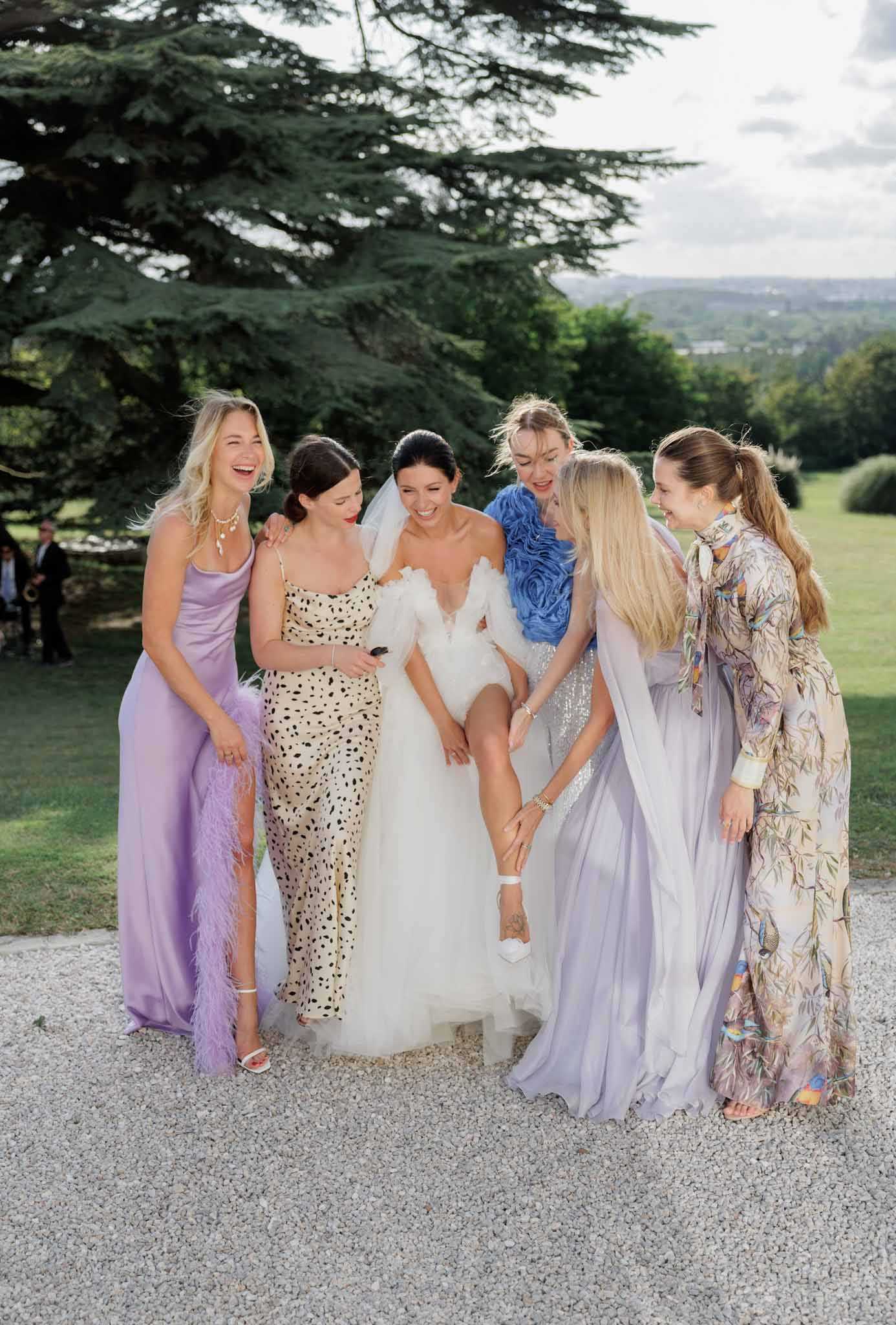 A bride and five bridesmaids pose together on a gravel path in the grounds of what appears to be a French chateau or estate, with manicured lawns and a large cedar tree visible in the background. The bride wears a white off-shoulder ball gown with a veil and white heeled sandals, and she has lifted her skirt to show one leg while a bridesmaid adjusts or admires her shoe, causing the group to laugh. The bridesmaids wear mismatched individual outfits rather than a uniform look: a lilac satin gown with feather trim, a cream and black dalmatian-print column dress, a blue feathered and ruffled long-sleeve garment, a periwinkle chiffon maxi dress with a flowing cape, and a long-sleeve floral-print maxi dress in muted lavender and earth tones. The shot is a full-length portrait taken at a slight distance, capturing the candid, joyful interaction among the group.