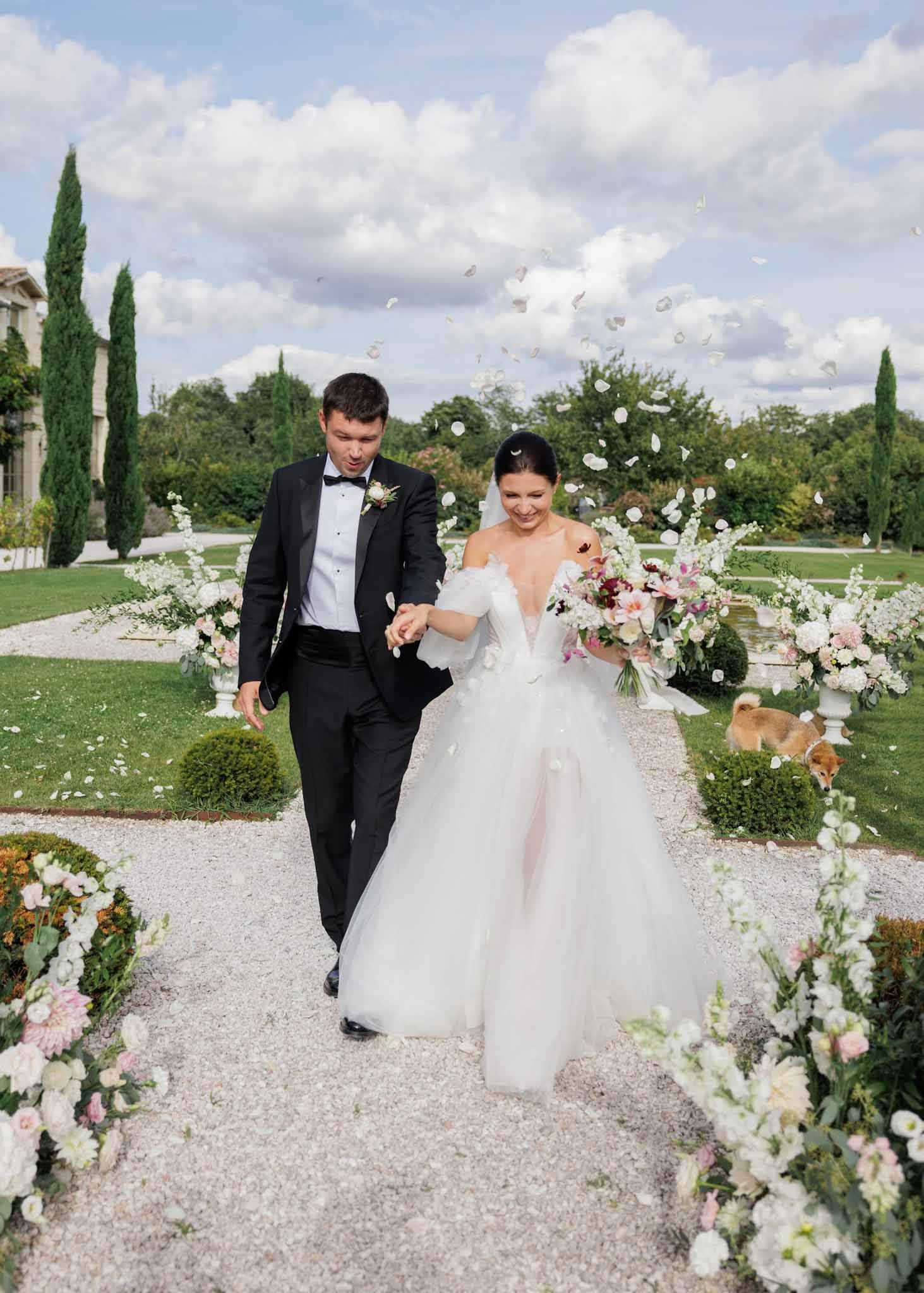 A couple walks hand-in-hand down a gravel garden path immediately following their outdoor ceremony, with white and blush flower petals being tossed into the air around them. The bride wears a white off-the-shoulder ballgown with a full tulle skirt and holds a lush bouquet mixing burgundy, blush, coral, and white blooms with greenery, while the groom wears a black tuxedo with a white boutonniere featuring small white flowers. The path is lined on both sides with abundant floral arrangements in white, blush pink, and pale peach — including what appear to be white delphinium, ivory roses, blush dahlias, and eucalyptus — displayed in white urn-style pedestals and low ground arrangements. A small shiba inu dog is visible in the background to the right, and the formal garden setting features manicured topiary, cypress trees, and a stone manor house partially visible at the rear left, suggesting a classic French or Italian-style estate venue. Wide portrait shot taken at ground level.