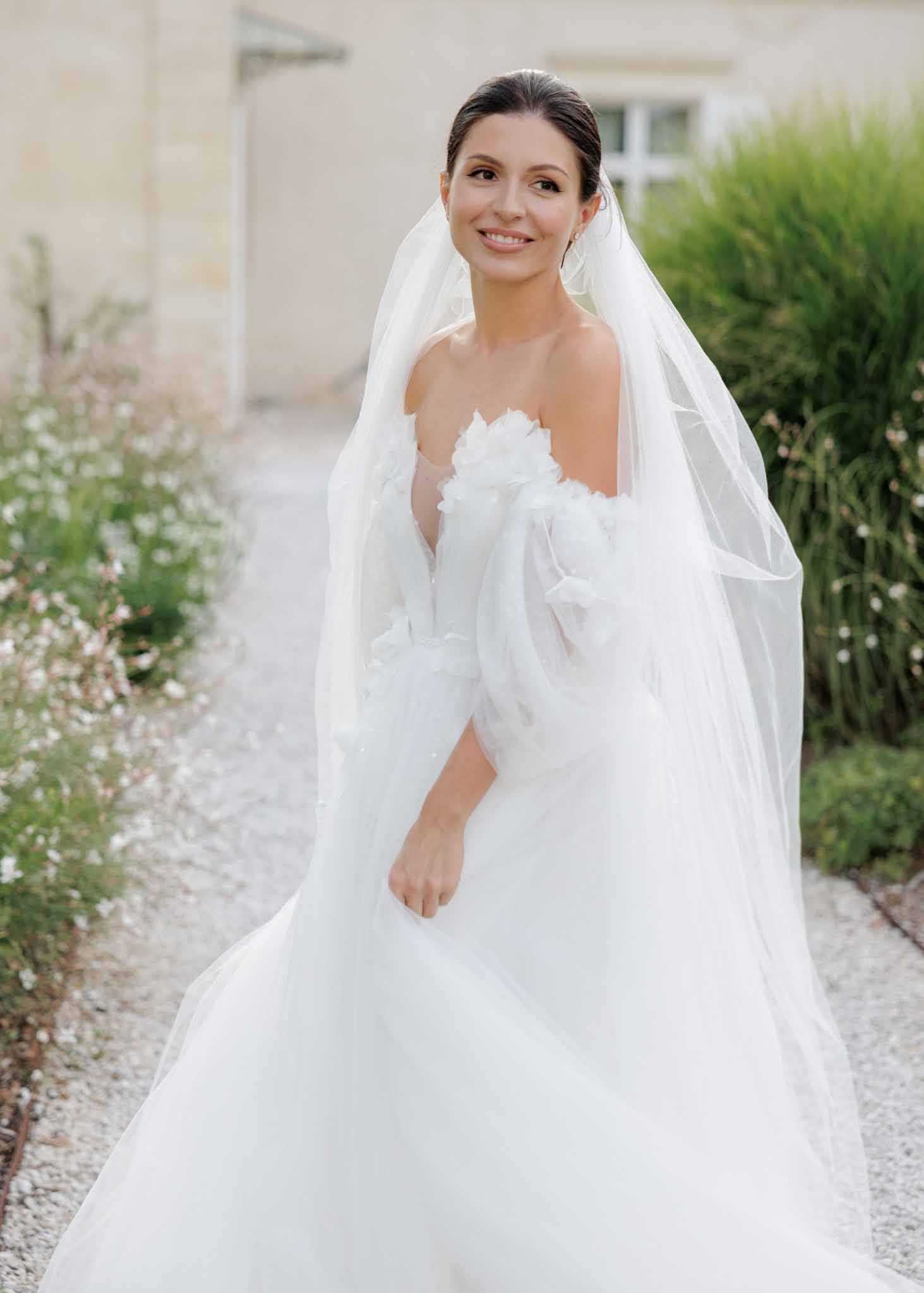A bridal portrait taken outdoors on a gravel path beside a French chateau or manor house. The bride, a dark-haired woman with her hair pulled back sleekly, wears a white tulle ball gown with off-the-shoulder puff sleeves and three-dimensional floral appliqués at the bodice, paired with a long cathedral-length veil. She faces the camera with a relaxed smile, one hand gently held at her side. The styling is classic and refined, with a modern romantic feel. The shot is a mid-length portrait with soft, even natural light.