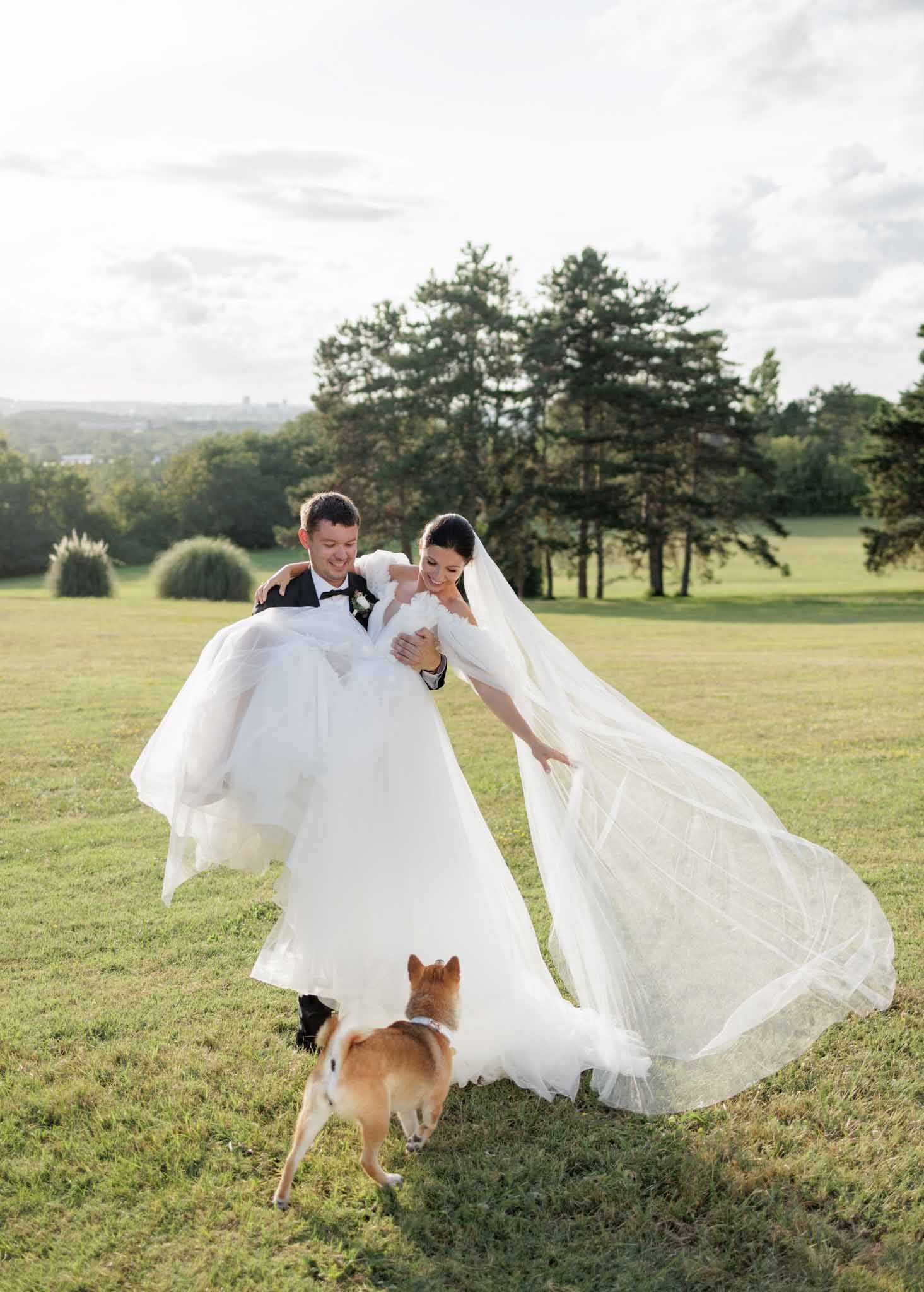 Outdoor couple portrait taken on a wide open lawn with a panoramic parkland backdrop. The groom, wearing a black tuxedo with a black bow tie and a small white boutonnière, is carrying the bride in his arms; both are laughing and looking downward. The bride wears a voluminous white ballgown with an off-the-shoulder neckline and a very long cathedral-length veil that billows outward in the wind. A small tan Shiba Inu dog wearing a white collar or bandana walks at their feet, appearing to look up at them. The overall styling is classic and formal. Full-length wide shot with warm late-afternoon light.