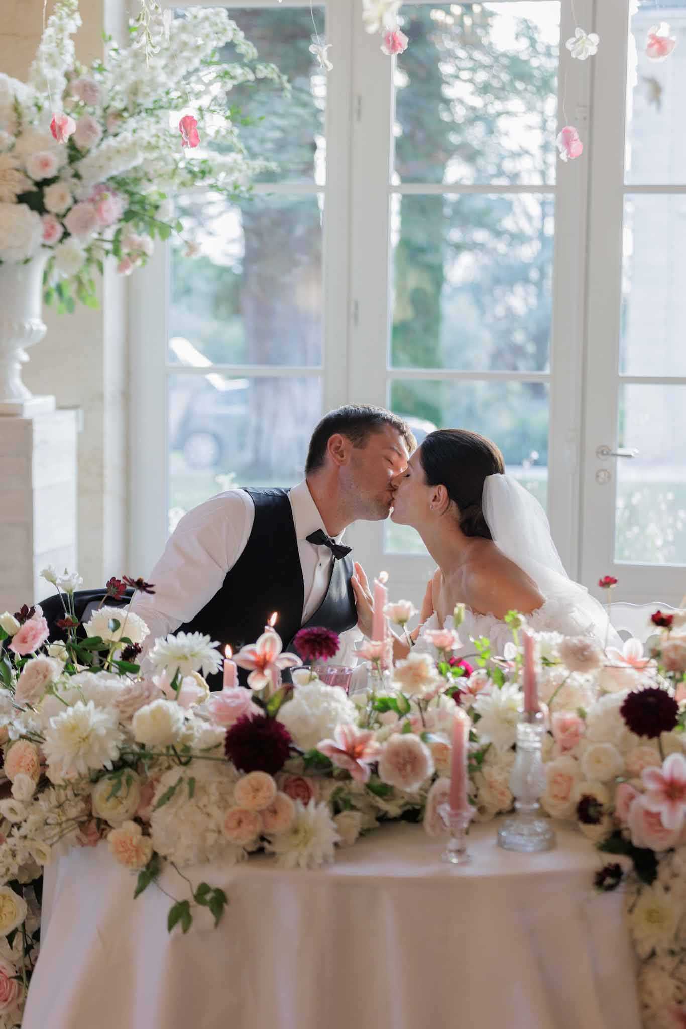 The bride and groom share a kiss while seated at their sweetheart table during the wedding reception, set inside a bright room with tall French doors overlooking greenery. The groom wears a white shirt, dark vest, and black bow tie, while the bride wears a white strapless dress with a veil and has dark hair styled up. The sweetheart table is densely covered in a lush floral runner featuring ivory garden roses, cream dahlias, burgundy dahlias, blush garden roses, peach roses, and pink Oriental lilies, with lit blush taper candles and small glass bud vases interspersed throughout. Overhead, suspended floral installations with blush and hot pink blooms and white flowers add vertical decor, and a large white floral arrangement on a column pedestal is visible to the left. The overall decor palette is romantic and rich, combining ivory, blush, peach, and deep burgundy tones in a classic indoor reception style. The shot is a medium portrait framing the couple centrally behind the floral-filled table.