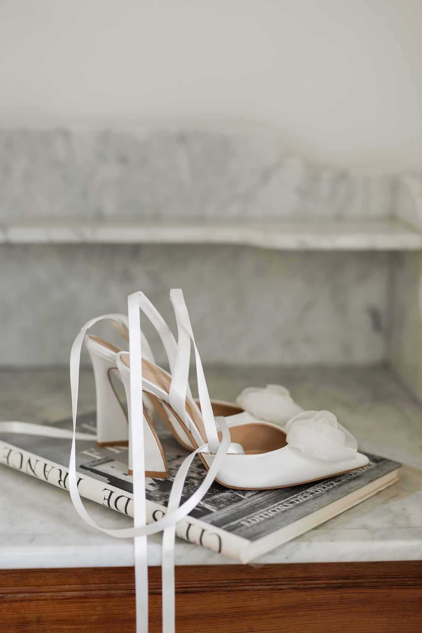 A detail close-up shot of two bridal shoes placed on top of a French-language coffee table book resting on a marble surface. One shoe is a white satin stiletto heel with long white ribbon ankle ties cascading down, while the other is a white flat or low-heeled shoe adorned with a large white organza flower at the toe. The tan leather insoles are visible on both shoes. The styling is clean and minimal, with the white shoes contrasting against the dark cover of the book, which appears to read 'De France' on its spine. The marble surface and background contribute to an all-white, modern aesthetic.