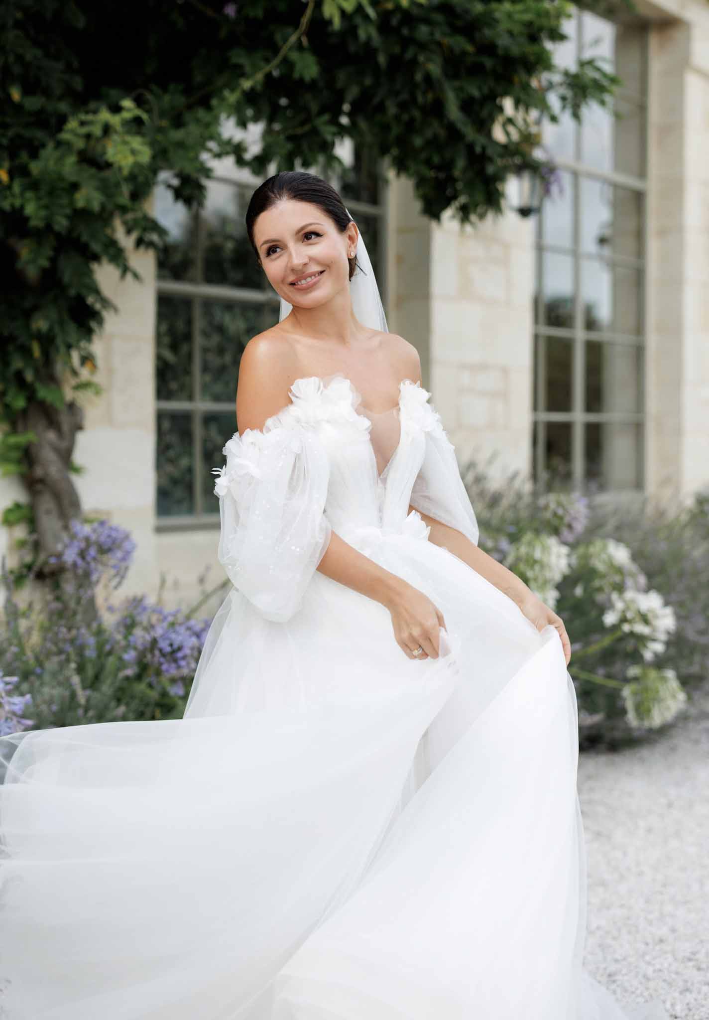 A bridal portrait taken outdoors in front of a French limestone building with large grid-pane windows and climbing foliage. The bride is seated, smiling off-camera, wearing a white off-the-shoulder ball gown with sheer puff sleeves, a deep V-neckline, and three-dimensional white floral appliqués along the neckline and sleeves; the full tulle skirt spreads widely in the foreground. She wears a simple white veil, her dark hair pulled back smoothly, and a solitaire engagement ring is visible on her hand. Lavender and white flowering plants are visible in the soft-focus background, contributing a romantic garden setting. The composition is a mid-length portrait shot with a shallow depth of field, styled in a modern classic aesthetic.