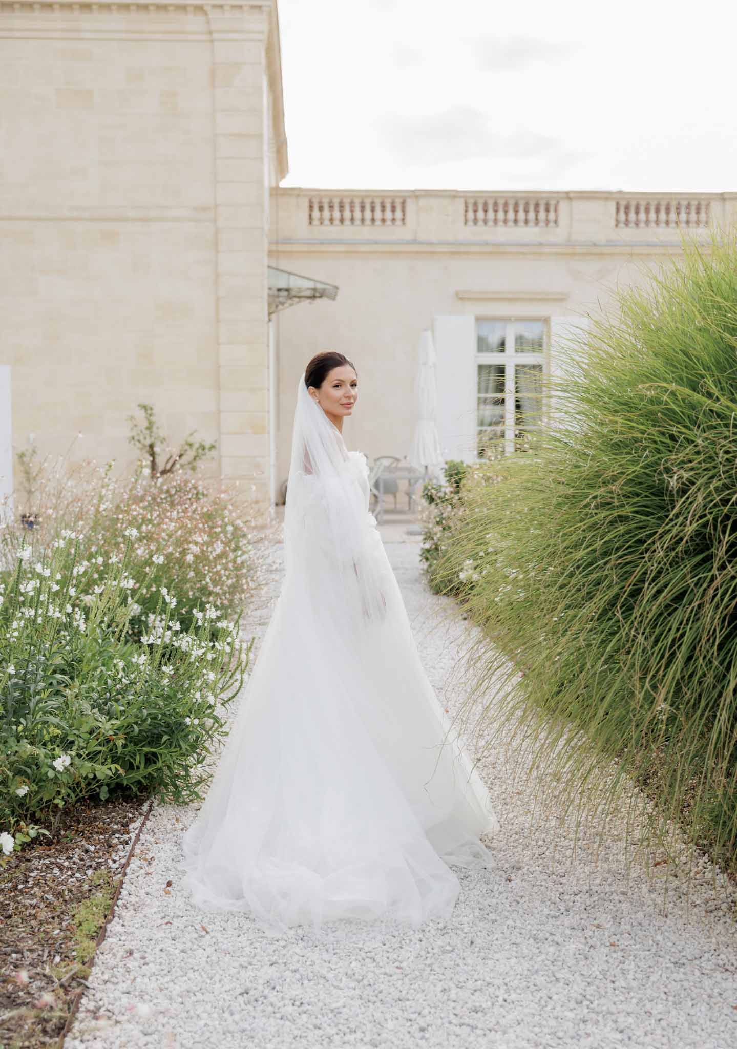 A bridal portrait taken outdoors on a white gravel pathway alongside a French chateau. The bride stands facing away from the camera and glances back over her shoulder, wearing a full-length white tulle ball gown with a long cathedral-length veil draped over her shoulders and trailing behind her. Her dark hair is styled in a low updo. The pathway is flanked by garden beds with small white blooms and tall ornamental grasses, with the pale limestone facade of the chateau visible in the background. The composition is a full-length portrait shot with a soft, airy light treatment consistent with an overcast day.