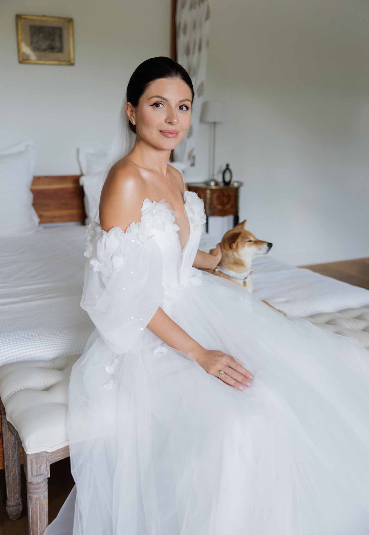 A bride sits on the edge of a bed in an indoor bedroom setting, posed in a three-quarter portrait shot. She wears a white off-the-shoulder ball gown with three-dimensional floral appliqués along the bodice and sheer tulle sleeves with scattered sequin details, paired with a veil and pearl stud earrings; her dark hair is pulled back into a sleek low bun. She rests one hand gently on a Shiba Inu dog sitting beside her on the bed, with an engagement ring visible on her finger. The room features antique wood furniture including a carved bedside table, white bedding, and a framed painting on the wall, suggesting a classic chateau-style interior.
