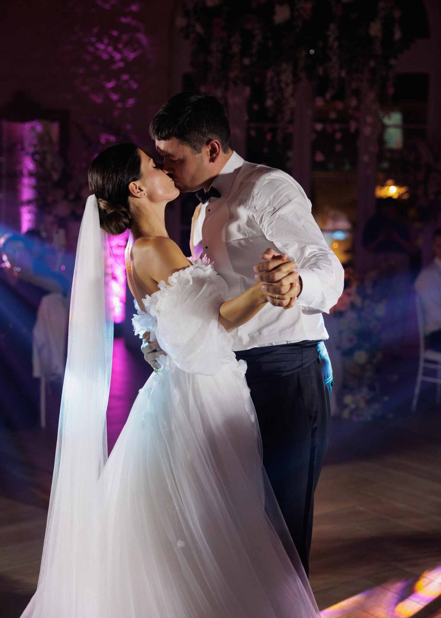 The bride and groom share a kiss during their first dance on a parquet dance floor inside a reception venue. The bride wears an off-the-shoulder white tulle ballgown with three-dimensional floral appliqué detailing on the bodice and long white gloves, with her dark hair styled in a low chignon and a long cathedral-length veil. The groom is dressed in a white dress shirt with a black bow tie and navy trousers, having removed his jacket. The indoor venue is lit with magenta and purple uplighting, with what appears to be hanging floral or botanical installations visible in the background, and seated guests are softly blurred in the background. The image is a mid-length portrait shot with the couple in sharp focus against the colorful, atmospheric reception lighting.