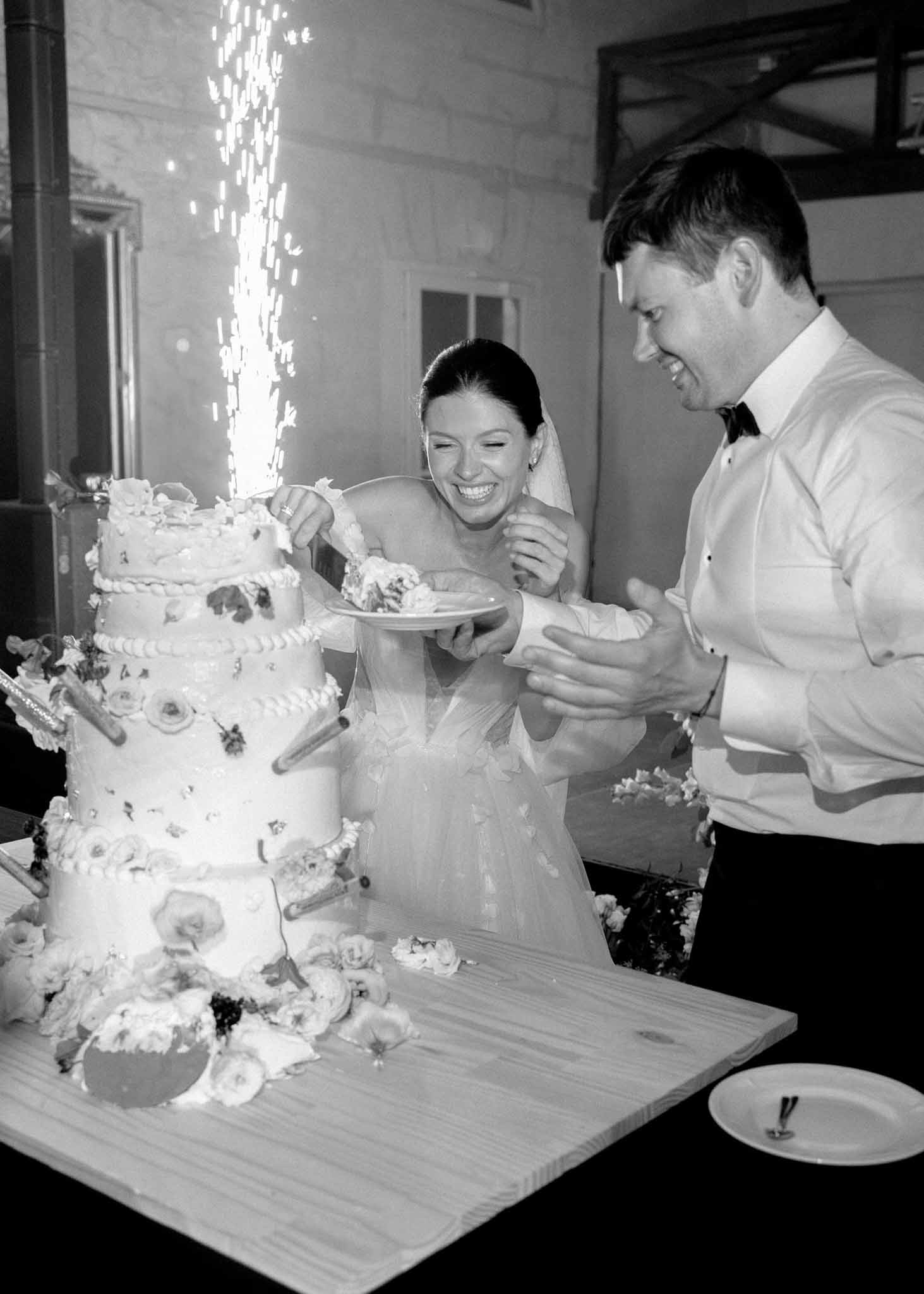 Black-and-white image of a bride and groom cutting their wedding cake during an indoor reception, with a cold sparkler fountain firing from the top tier of the cake. The bride, wearing a strapless gown and veil with her dark hair pulled back, is laughing and holding a plate with a slice of cake, while the groom in a tuxedo shirt and bow tie leans in smiling. The four-tier cake is decorated with floral appliqués and small cylindrical decorations at each tier, displayed on a wooden board with loose flowers scattered around the base. The composition is a medium close-up portrait shot with high contrast tones typical of black-and-white film-style photography.