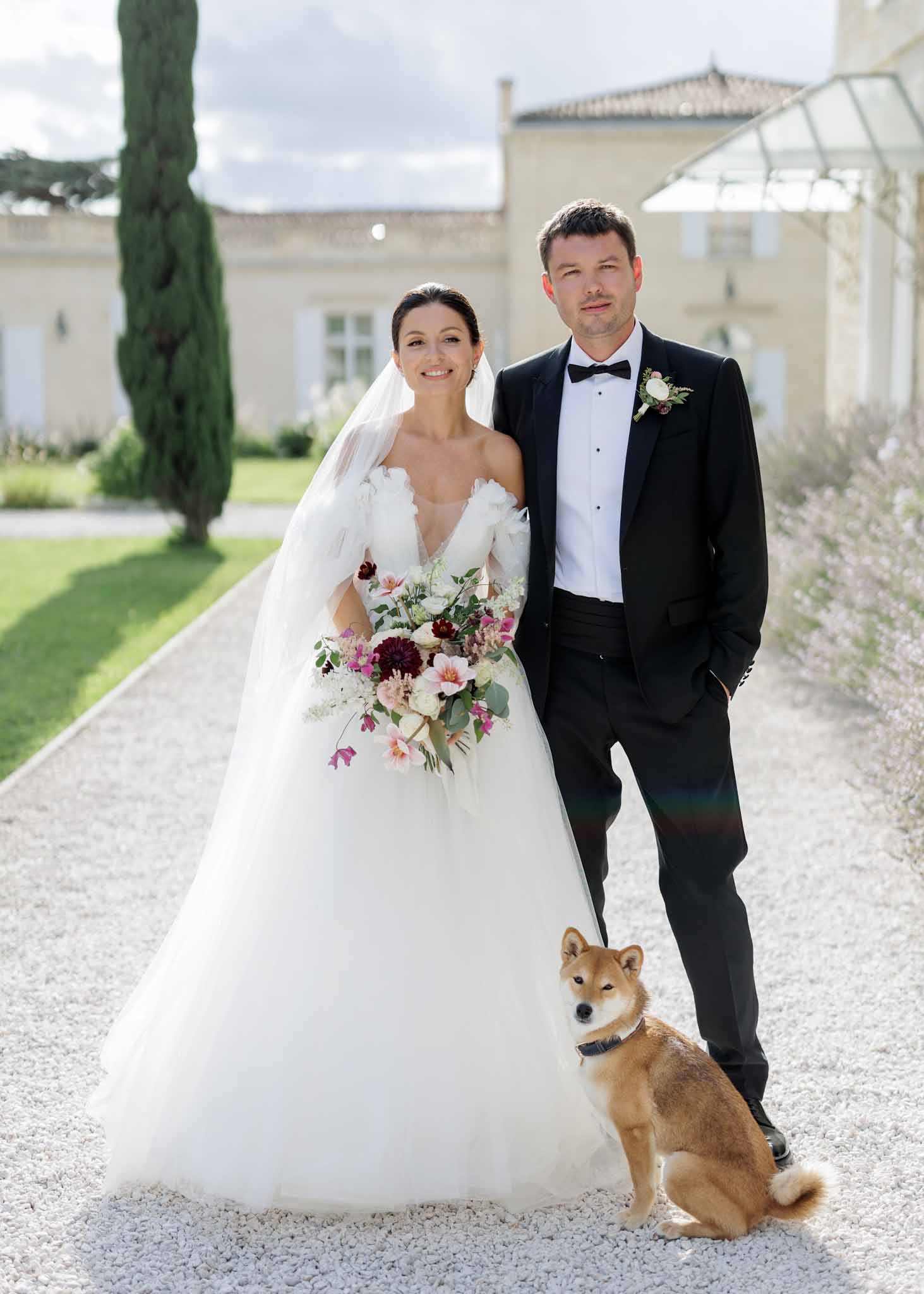 A couple portrait taken outdoors on a gravel driveway in front of a French chateau with stone façade and a glass greenhouse structure visible to the right. The bride wears a white tulle ballgown with an off-the-shoulder neckline featuring floral appliqué detailing and a cathedral-length veil, and holds a large cascading bouquet of burgundy dahlias, blush and pink blooms, white flowers, and trailing eucalyptus greenery. The groom wears a black tuxedo with bow tie, white dress shirt, and a boutonnière of white and burgundy flowers. A tan and white Shiba Inu dog sits at their feet. The portrait is a medium full-length shot with a classic, formal styling approach.