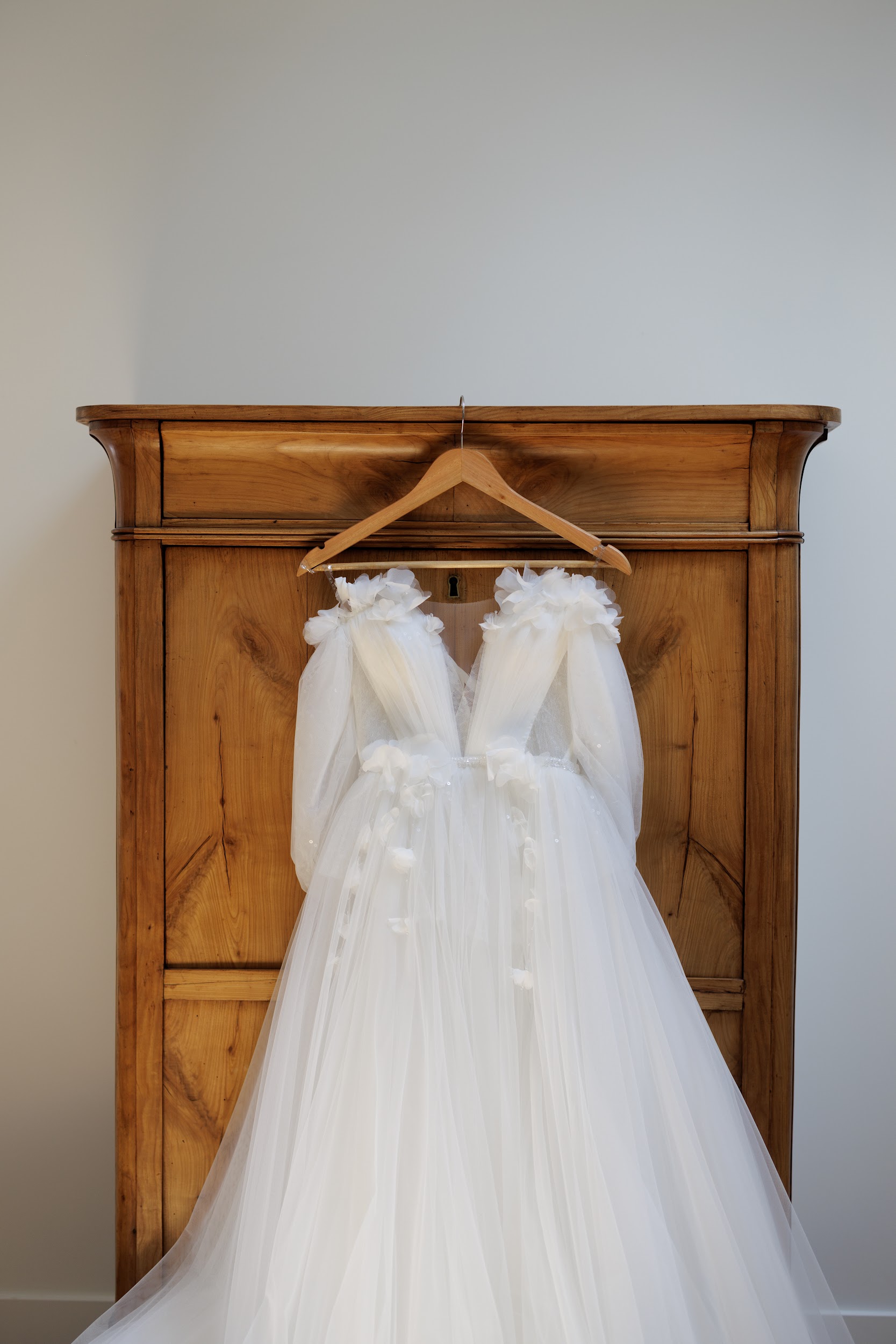 A getting-ready detail shot of a white bridal gown hanging on a wooden hanger hooked to an antique honey-toned wooden armoire. The dress features a deep V-neckline, sheer long tulle sleeves, 3D floral appliqués at the shoulders and waist, and a full layered tulle skirt that cascades to the floor. The image is a centered, medium-distance portrait-style composition with the armoire set against a plain light grey wall, giving the shot a clean, minimal look.