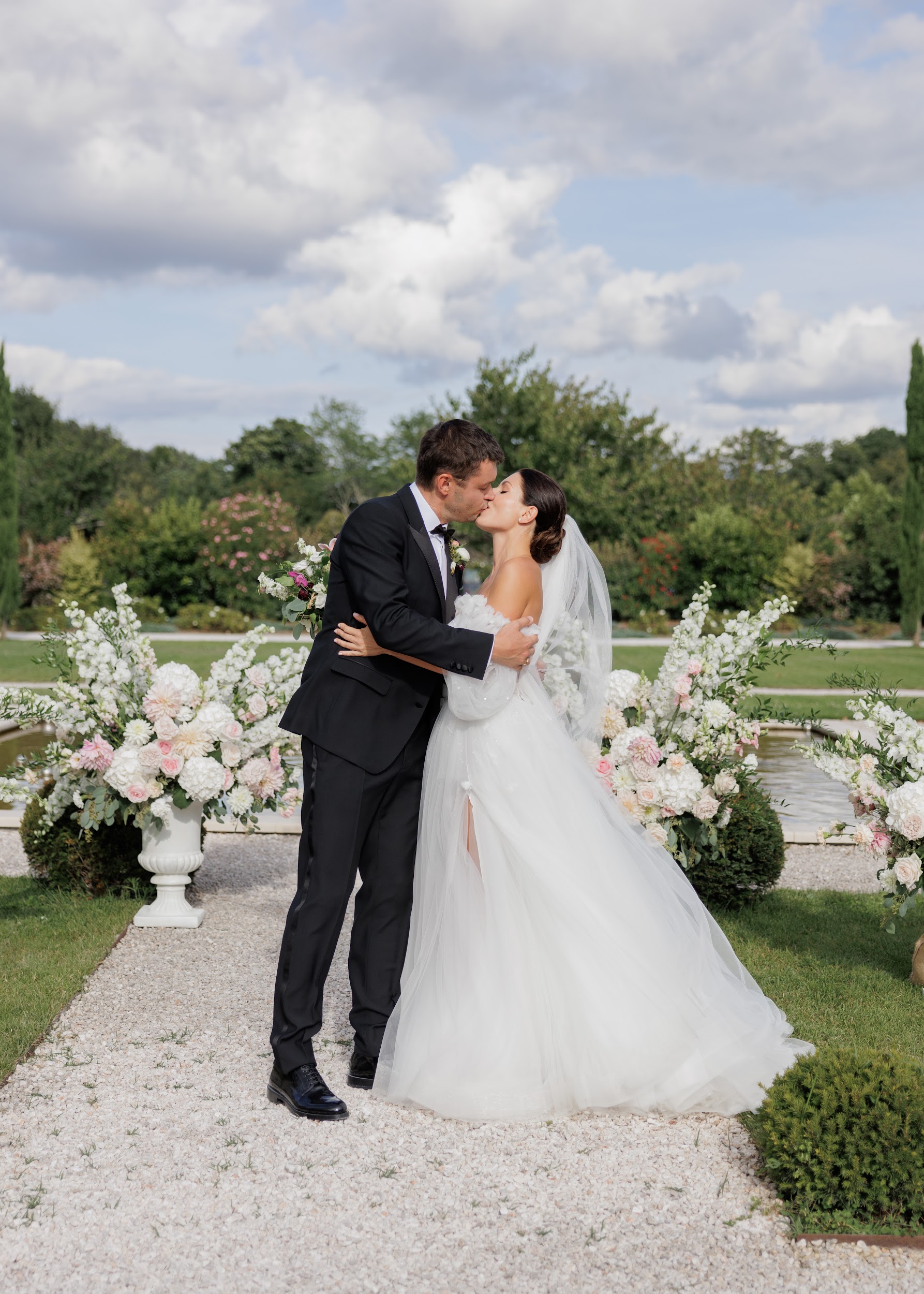 A couple shares a kiss during what appears to be an outdoor ceremony or portrait moment on a gravel path within formal French garden grounds, with a reflecting pool or water feature visible in the background. The bride wears a strapless white tulle ballgown with a cathedral-length veil, while the groom is dressed in a black tuxedo with a bow tie and boutonnière. Large white stone urns on either side of the couple are filled with full floral arrangements featuring white dahlias, blush pink roses, ivory garden roses, and trailing greenery, forming a natural floral aisle. The shot is a mid-length portrait composition capturing both figures from head to toe against the garden backdrop.