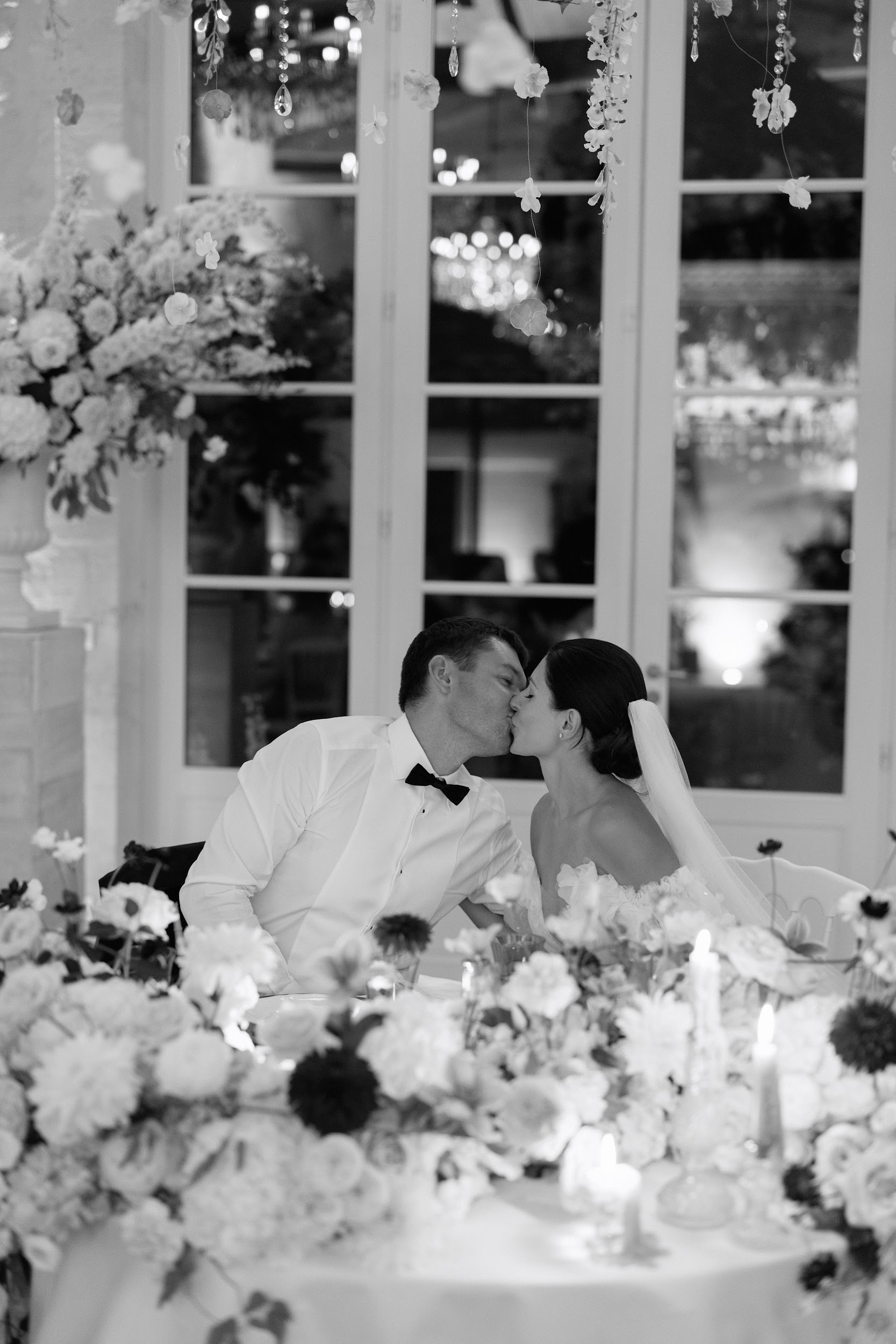This black-and-white image shows a bride and groom kissing at their sweetheart table during an indoor evening reception. The groom wears a white dress shirt with a black bow tie, and the bride wears a strapless gown with her dark hair styled up, with a veil trailing behind her. The foreground features a dense, low floral arrangement of dahlias, ranunculus, and mixed blooms with tall taper candles, rendered in deep contrasting tones. Overhead, crystal drop garlands and floral installations hang from above, and large French doors in the background reflect a chandelier and warm exterior uplighting, creating bright highlights against the dark night. The composition is a medium portrait shot taken slightly above table level, with the floral centerpiece in soft focus in the foreground.
