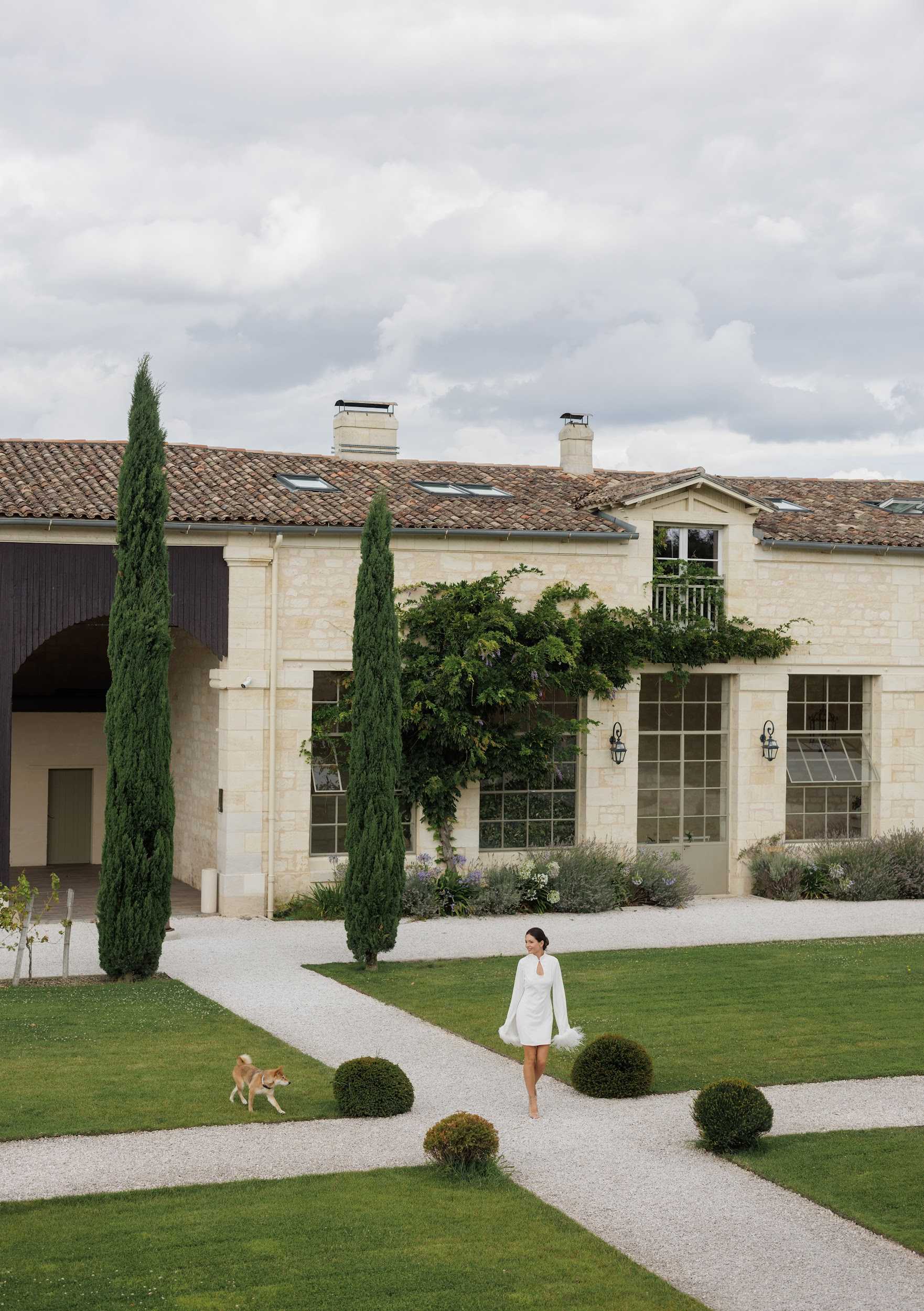 A bride wearing a short white long-sleeved mini dress with flared cuffs and a keyhole neckline walks barefoot along a gravel pathway in the manicured grounds of a French stone estate. A tan Shiba Inu dog walks alongside her on the lawn. The venue is a classic French limestone building with a terracotta tile roof, large steel-framed windows, climbing wisteria, and a small balcony with white railings, flanked by tall cypress trees and clipped box ball topiaries. The wide-angle portrait shot captures both the bride at mid-ground and the full facade of the building, giving prominence to the venue architecture and formal garden layout. Potential venue feature image.