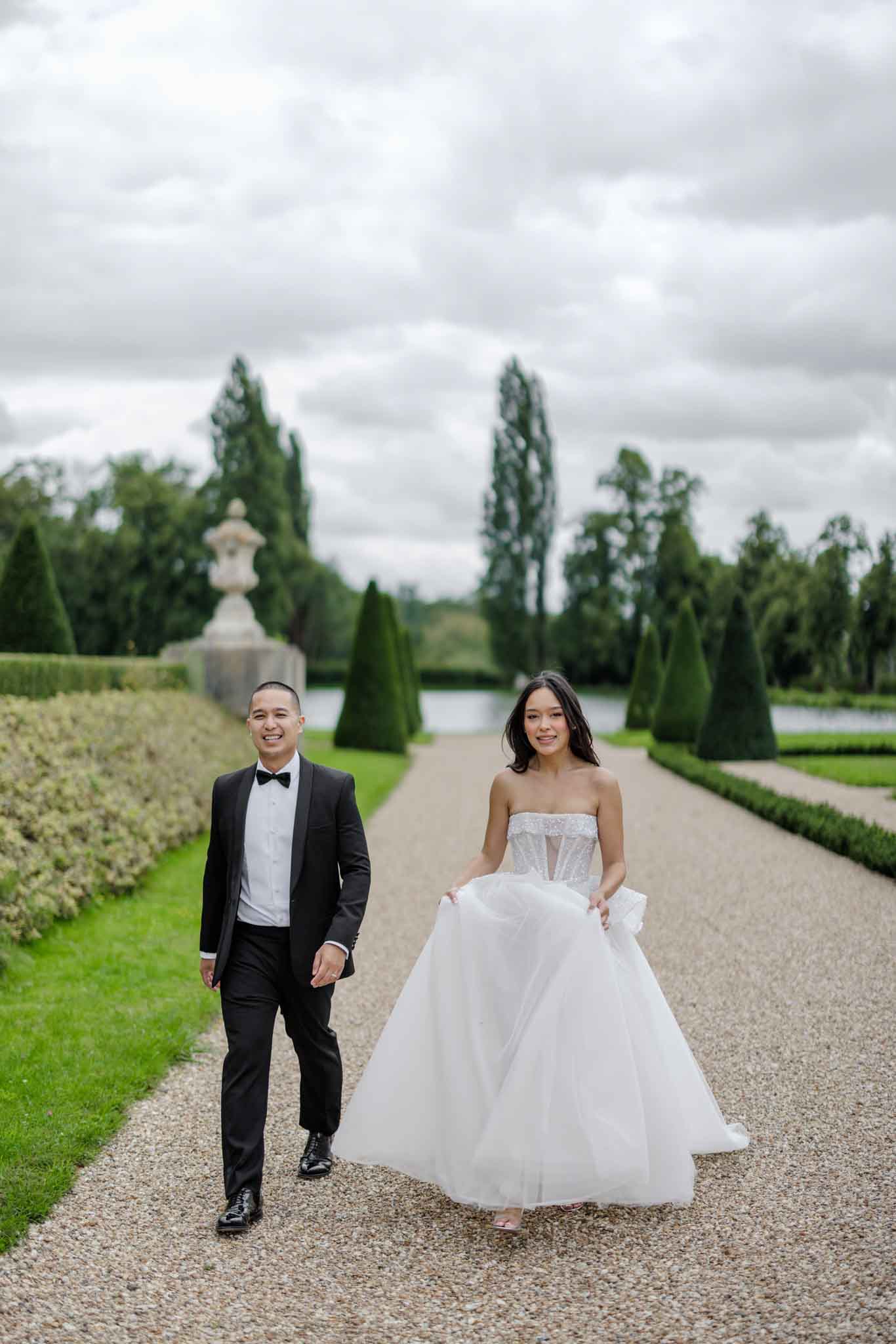 A couple walks together along a formal gravel garden path during what appears to be a portrait session. The groom wears a black tuxedo with a black bow tie and white dress shirt, while the bride wears a strapless white ballgown with a beaded corset bodice and a full tulle skirt, which she lifts slightly as she walks. The outdoor setting features a formal French garden with neatly trimmed conical topiary trees, sculpted hedgerows, a stone urn pedestal, and a reflecting pool or canal visible in the background, consistent with a chateau or grand estate garden. The composition is a full-length portrait shot with the couple positioned centrally, and the formal garden's symmetrical layout receding into the background.