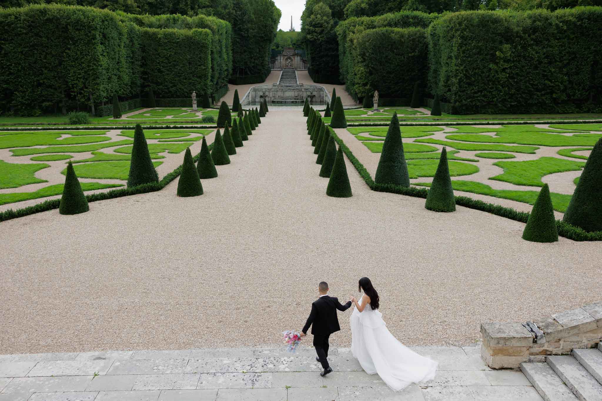 A couple portrait taken from an elevated angle, showing a bride and groom walking hand-in-hand along a grand gravel allée in a formal French-style garden. The groom wears a black suit and the bride wears a white full-length gown with a flowing train, carrying a bouquet with pink and colorful blooms. The garden features precisely clipped conical topiary trees lining the central path, symmetrical parterres with curved boxwood hedging, and a stone staircase with a decorative fountain or ornamental feature at the far end. The wide-angle, near-aerial composition emphasizes the strong linear perspective of the garden's formal design, with the couple positioned small in the frame at the foreground. Potential venue feature image.