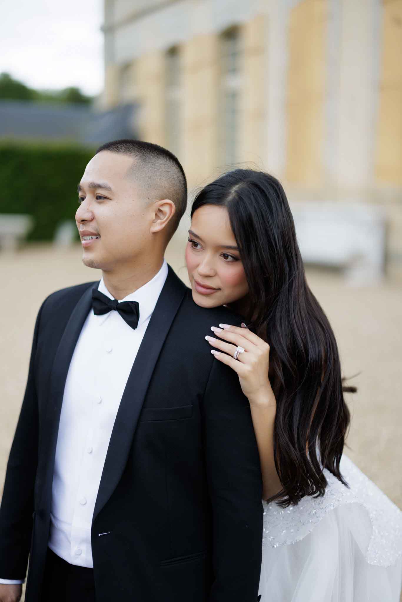 A couple portrait taken outdoors in front of a large classical French chateau with pale yellow stone facade visible in the blurred background. The groom wears a black tuxedo with a black bow tie and white dress shirt, while the bride, dressed in a white gown with visible beaded or lace detailing at the shoulder, rests her head against his back and places her hand on his shoulder, displaying a diamond engagement ring and wedding band with white-tipped manicured nails. The groom looks off to one side while the bride faces the camera directly. The styling is classic and formal, with a black-and-white color palette for the couple's attire. The shot is a close-up portrait with shallow depth of field, keeping the architectural background softly out of focus.