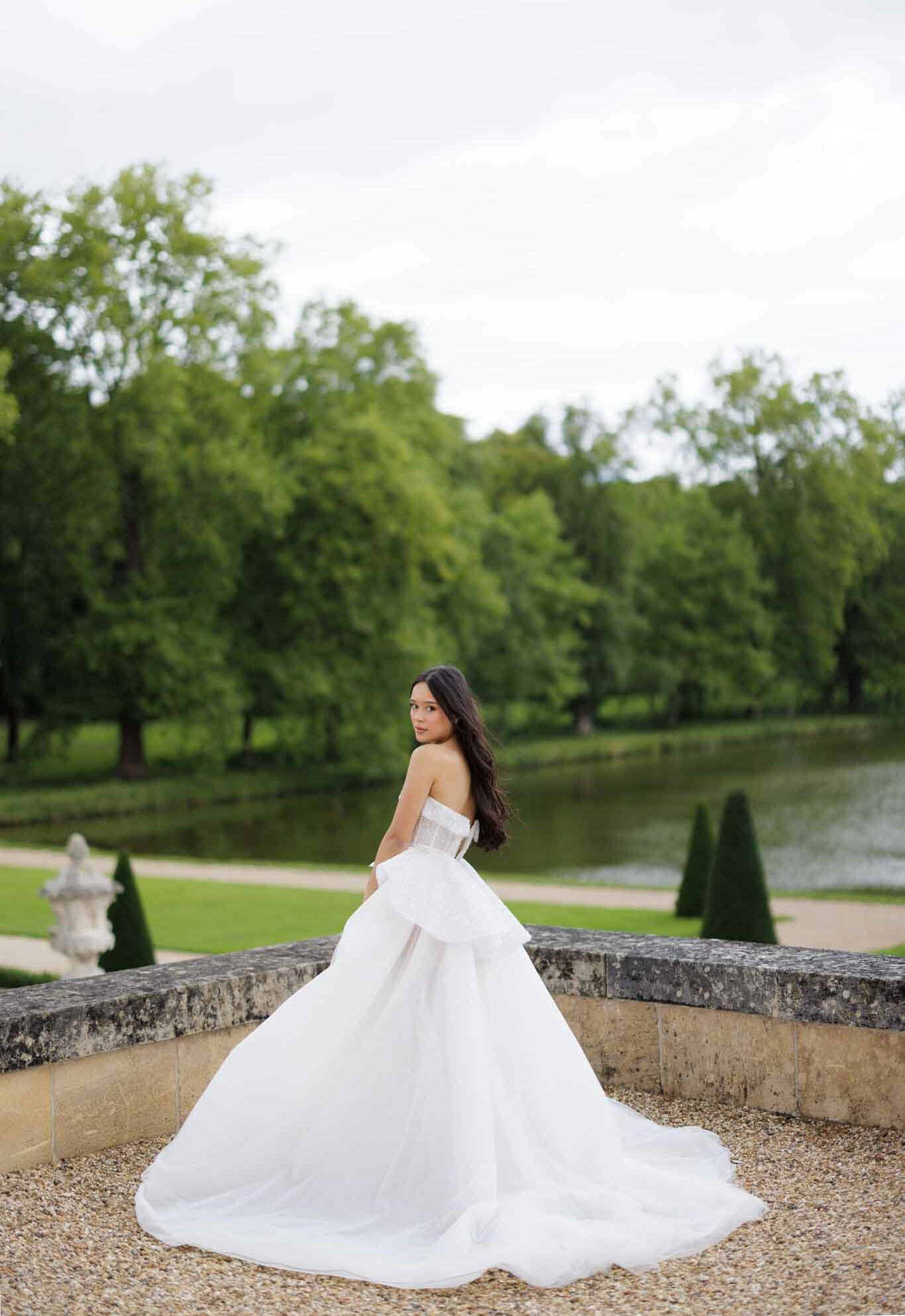 A bridal portrait taken outdoors on a gravel terrace of a formal French chateau garden, with a bride posing from behind and glancing back over her shoulder toward the camera. She wears a white strapless ball gown with a structured corseted bodice featuring subtle embellishment, a voluminous layered skirt, and an extended cathedral-length train spread across the gravel. Her long dark hair falls loosely down her back. The formal garden behind her features manicured conical topiary hedges, a stone balustrade, a classical urn sculpture, a reflecting pool or ornamental lake, and a manicured lawn, consistent with a classic French chateau estate. The composition is a full-length portrait shot from a low-to-mid angle, emphasizing the sweep of the train in the foreground. Potential venue feature image.