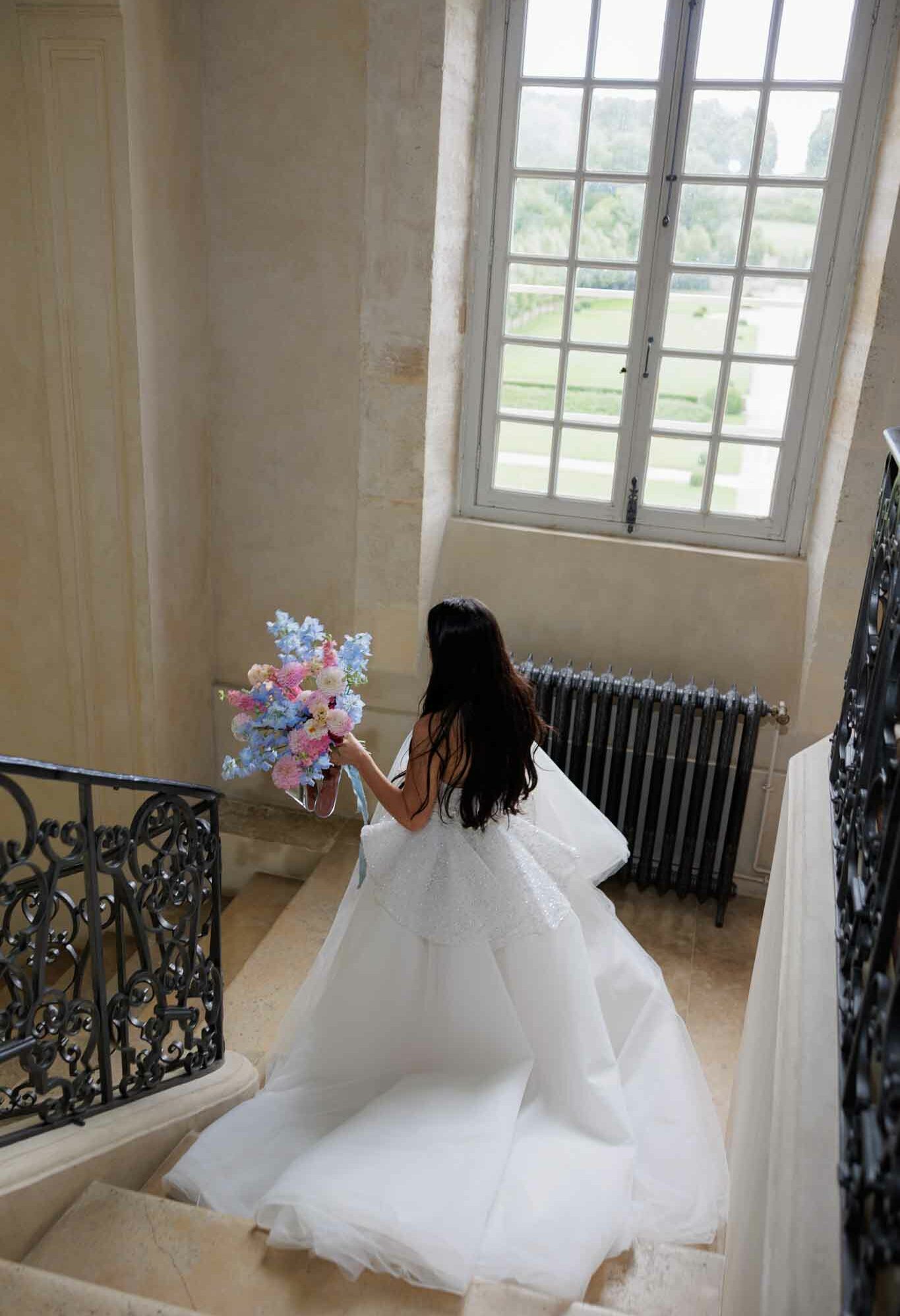 A bride with long dark hair is photographed from above as she descends a stone staircase inside a chateau, shot from an elevated angle looking down. She wears a voluminous white ball gown with a full tulle skirt and what appears to be an open or low back, with a substantial train spreading across the stairs. She holds a large, loose bouquet of blue delphiniums, pink dahlias or cosmos, and white blooms with trailing pale blue ribbons. The interior features ornate wrought-iron stair railings, warm limestone walls and steps, a cast-iron radiator, and tall French windows with gridded panes overlooking formal green grounds. The overall styling is classic and romantic with a soft pastel floral palette contrasting against the white gown.