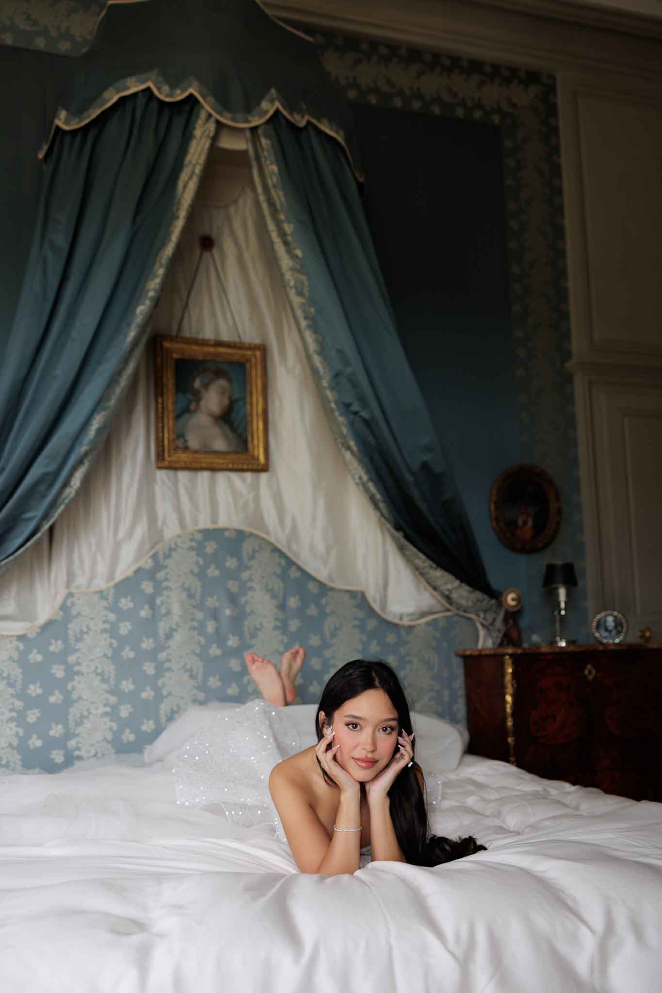 A bride is lying on a large canopied bed during a getting-ready or portrait session inside a chateau bedroom. She is wearing a white sequined or beaded strapless gown and a delicate pearl or diamond bracelet, with long dark hair worn loose. The room features an ornate teal and ivory silk canopy draped above the bed, blue-and-white toile upholstered headboard, dark wood antique bedside furniture with gilded detailing, and a gold-framed oil portrait painting hanging from the canopy. The walls are covered in blue floral wallpaper, and a small oval portrait and a miniature lamp are visible on the nightstand. The composition is a medium portrait shot taken from the foot of the bed, with the bride resting her chin on her hands and looking directly at the camera. The overall decor style is French classical or 18th-century aristocratic.