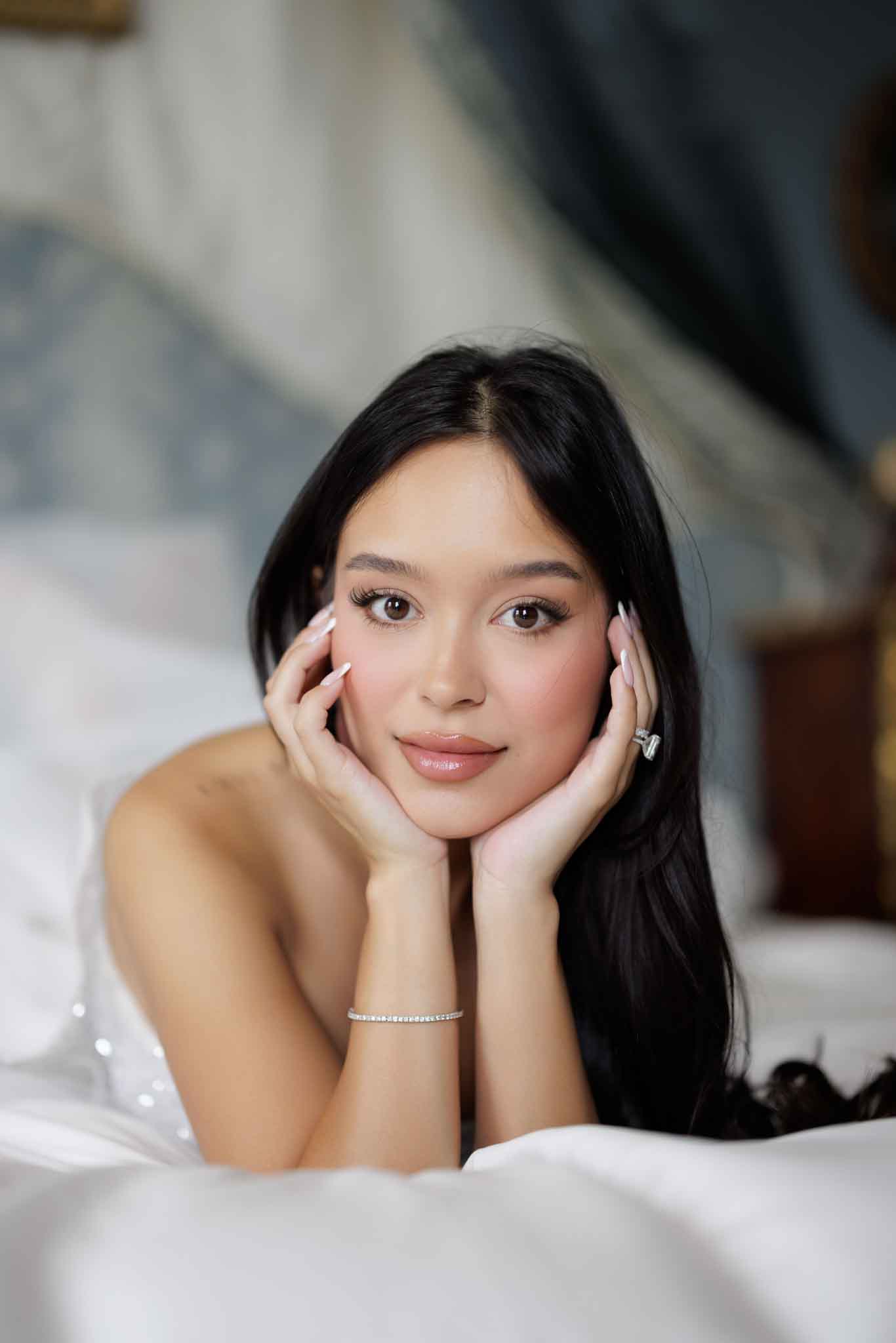 A close-up portrait of a bride lying on a white surface, resting her face in both hands and looking directly at the camera. She has long dark hair worn loose, a natural glam makeup look with defined lashes and a neutral lip, white almond-shaped nails, a diamond tennis bracelet, and a diamond ring visible on her right hand. She is wearing a strapless white gown with subtle embellishment along the bodice edge. The background is softly blurred but suggests an ornate interior room with pale blue decorative wall panels, consistent with a classic chateau or historic venue setting.