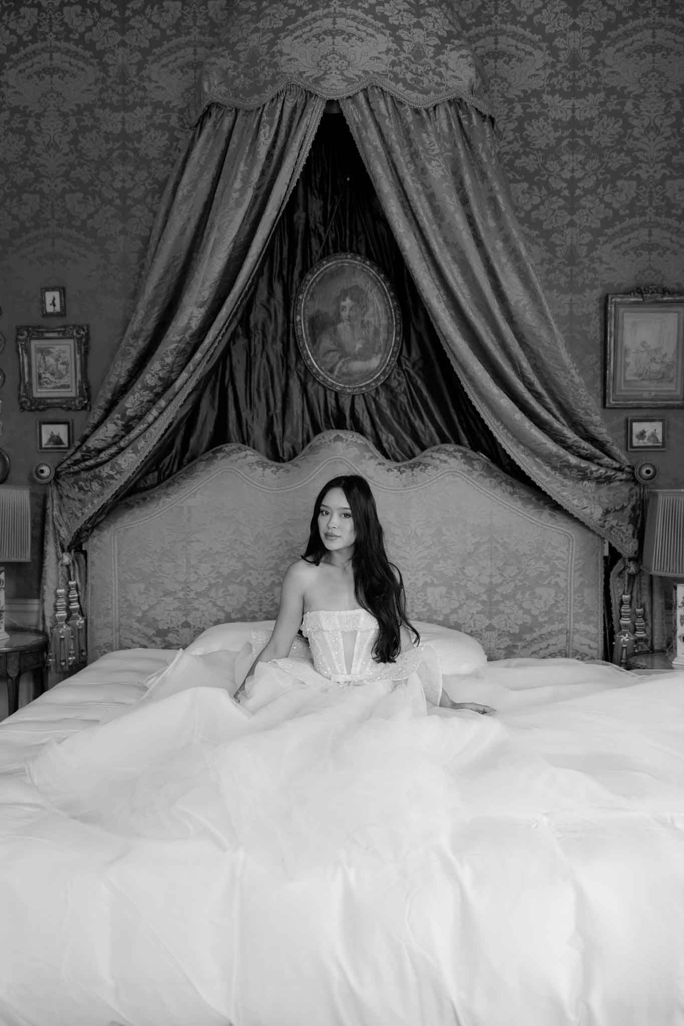 A black-and-white bridal portrait of a bride seated on a large ornate bed inside what appears to be a historic chateau or castle bedroom. She wears an off-the-shoulder corset-bodice ball gown with a voluminous tulle skirt that spreads widely across the bed, and her long dark hair falls loosely over her shoulders. The room features heavily draped fabric canopy above the upholstered headboard, damask-patterned wallpaper, multiple framed paintings and portraits on the walls, and bedside lamps on gilded side tables, giving the space a grand, classical French interior aesthetic. The image is a medium-distance portrait shot with high contrast, the bride centered against the dramatic canopy and an oval portrait painting framed within the drapes behind her.