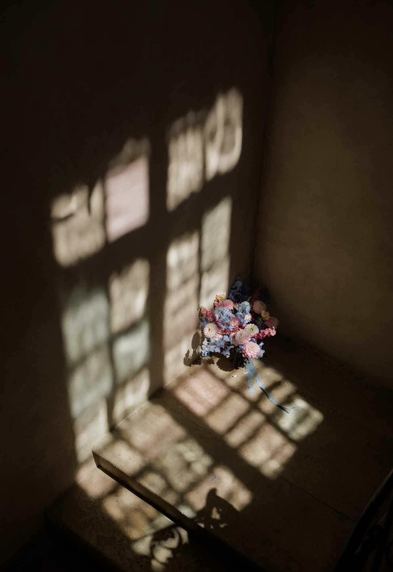 A detail shot of a bridal bouquet placed on a wooden surface, likely a windowsill or bench, in a dimly lit interior room. The bouquet features pink dahlias, pale blue hydrangeas, and small pink and white button flowers, tied with a trailing pale blue ribbon. Strong geometric window light casts a grid-like shadow pattern across the wall and surface, illuminating the bouquet against the otherwise dark surroundings. The composition is a medium overhead angle, emphasizing the interplay between the delicate pastel floral arrangement and the dramatic natural light and shadow.