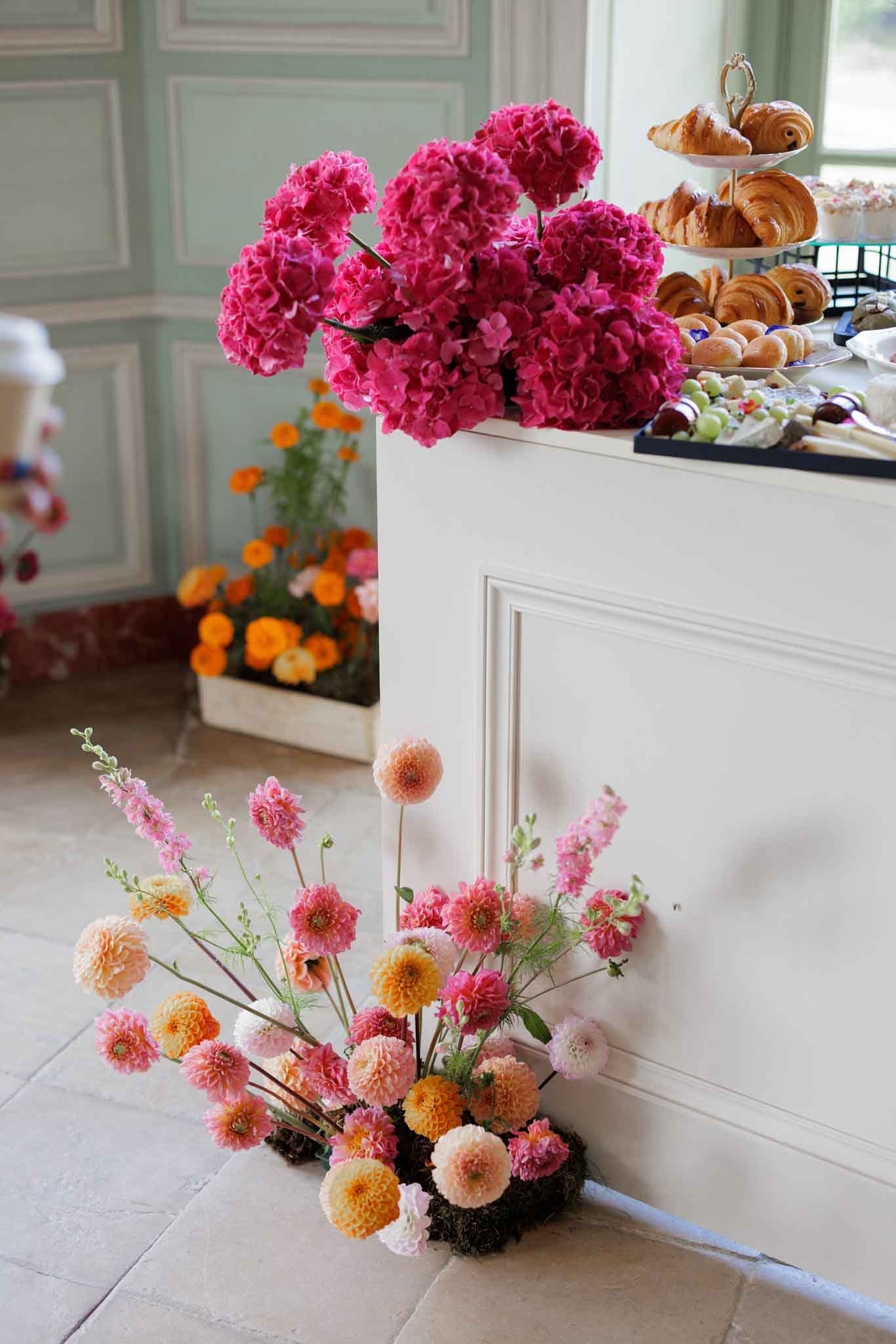 A detail shot of a catering or welcome table setup in an indoor venue with sage green paneled walls and stone tile flooring. In the foreground, a low floral arrangement sits directly on the floor, featuring pink, peach, and yellow dahlias, pink larkspur, and ranunculus set in a moss base. On top of the white paneled counter, a large arrangement of deep magenta hydrangeas spills dramatically over the edge, while a tiered stand holds croissants and pastries alongside trays of additional baked goods, fruit skewers, and bite-sized treats. A crate of orange and yellow marigolds is visible in the background. The overall floral palette spans hot pink, magenta, peach, orange, and soft pink, creating a bold, garden-style aesthetic.