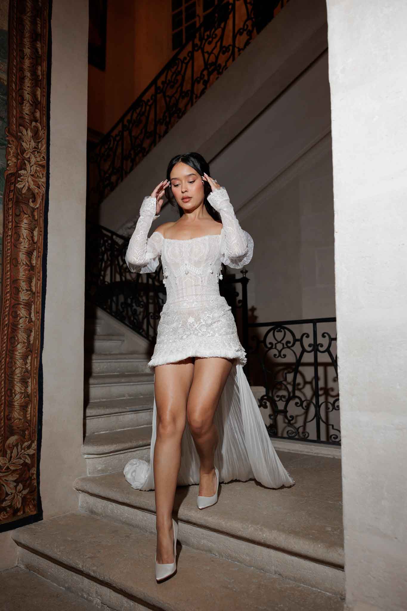 A bride poses alone on an indoor stone staircase featuring ornate wrought-iron railings, consistent with a chateau or historic manor interior. She wears a white heavily embellished mini wedding dress with a structured corset bodice, intricate lace and beaded detailing, sheer long sleeves, a fringe hem, and a detachable floor-length tulle train trailing behind her. The look is completed with white pointed-toe heels and her dark hair is pulled back. The composition is a full-length portrait shot framed by a white pillar and a decorative tapestry visible on the left, with warm ambient interior lighting creating a moody contrast. The overall styling aesthetic is modern bridal with a fashion-forward, high-couture edge.