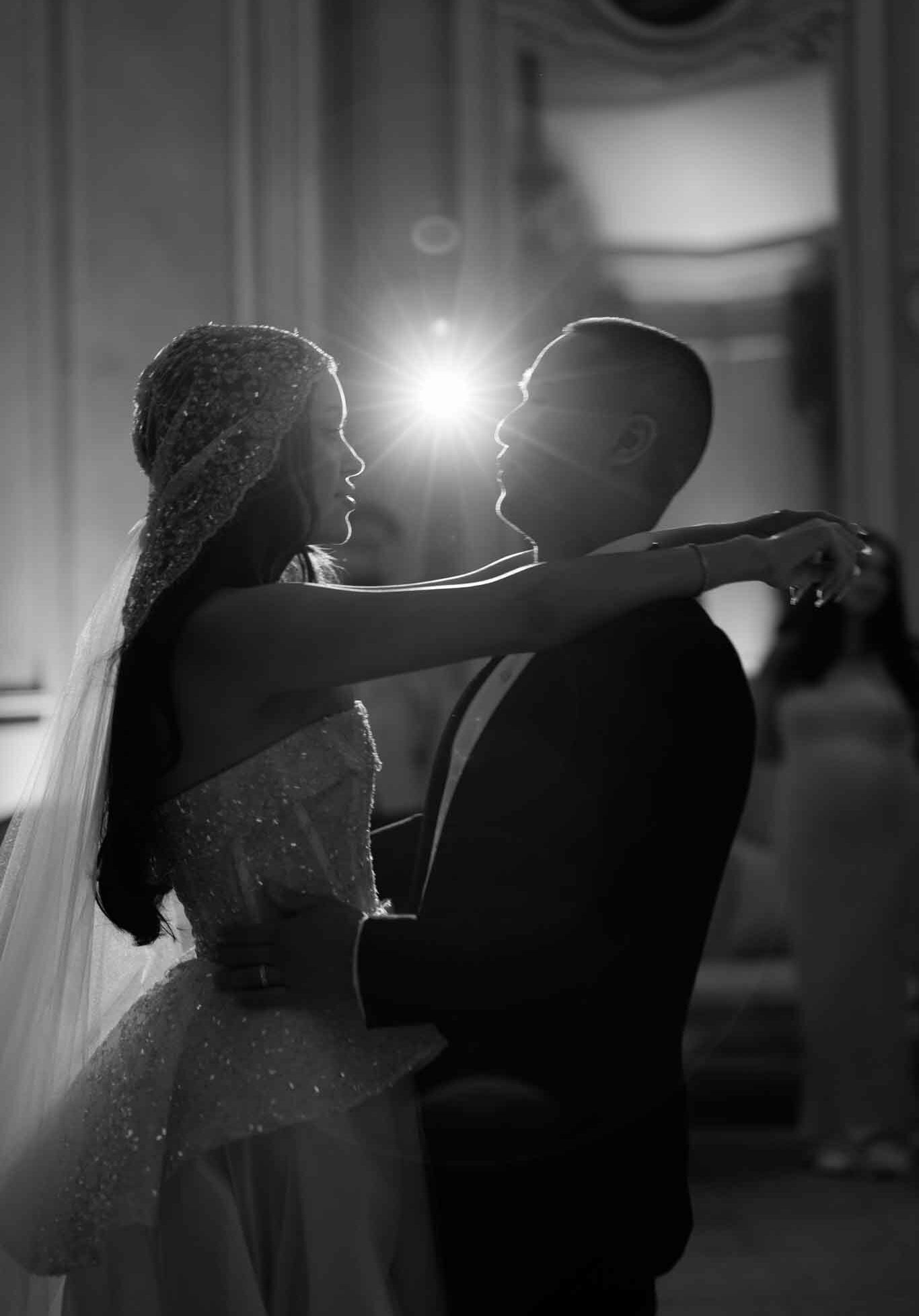 A black-and-white close-up portrait of a bride and groom sharing their first dance inside what appears to be a grand ballroom with ornate molded wall paneling visible in the background. The couple faces each other in an embrace, with the bride's arms draped around the groom's neck and his hands at her waist; a strong point-light source positioned directly between their faces creates a dramatic starburst lens flare effect. The bride wears a heavily beaded or embroidered strapless gown with a matching beaded capelet or hood over her head, along with a long veil; the groom is dressed in a dark suit. The image is rendered in deep blacks and bright highlights with high contrast, giving the silhouetted figures a dramatic quality. At least one blurred guest is visible in the background to the right.