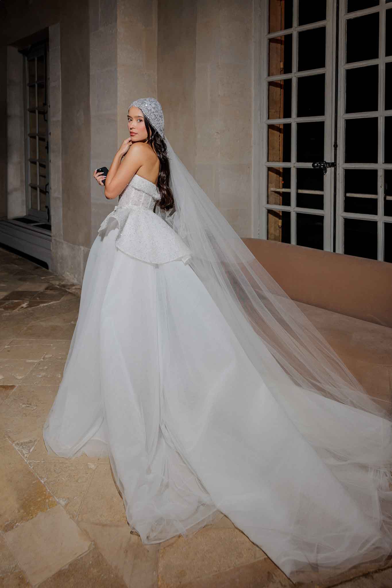 A bridal portrait of a single bride photographed outdoors against the stone facade of what appears to be a French chateau or historic building, with tall French doors visible in the background. The bride wears a strapless white ball gown featuring a heavily embellished lace and beaded corseted bodice with a structured peplum detail, paired with a voluminous soft tulle skirt with an extended cathedral-length train spread across the stone-paved floor. She accessorizes with a pearl and crystal-embellished juliet cap veil that extends into a full-length cathedral veil, and holds a small dark-colored clutch. The bride glances back over her shoulder toward the camera with long dark hair worn down. The shot is a three-quarter rear-facing portrait captured in natural and ambient light, with the stone architecture providing a classic, formal backdrop.