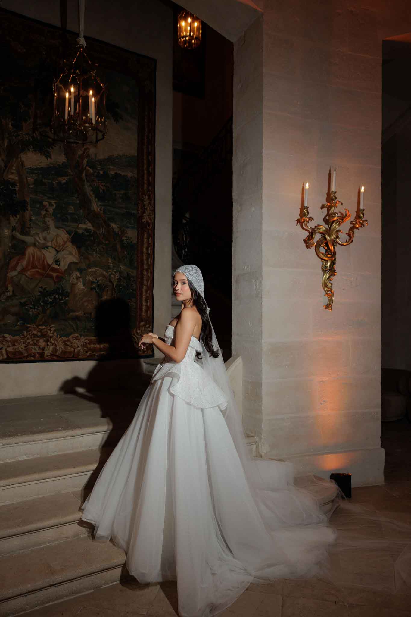 A solo bridal portrait taken indoors in what appears to be a French chateau, with the bride standing on stone steps and glancing back over her shoulder toward the camera. She wears a white strapless ball gown with a structured peplum bodice featuring textured or embroidered detailing, a full tulle skirt with a long train, and a lace or beaded juliet cap with a long veil trailing down her back. The setting is lit with warm amber uplighting, a gold ornate candelabra wall sconce with lit candles, and a hanging brass lantern with candle-style bulbs. A large antique tapestry with figurative scenes in earthy tones hangs on the wall to the left. The overall styling aesthetic is classic and formal with baroque-inspired decor. Full-length portrait shot with strong directional lighting creating dramatic shadows.