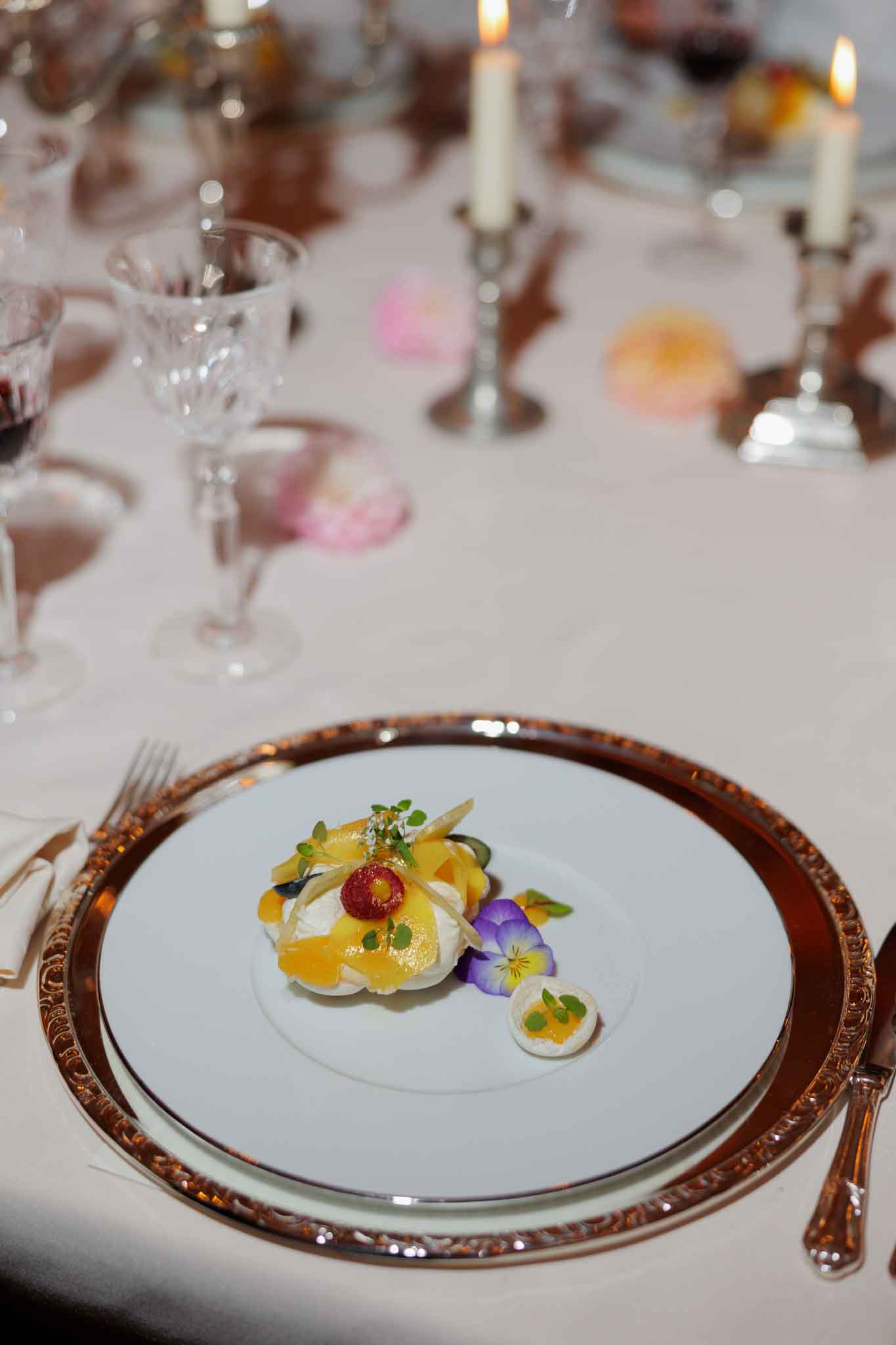 Close-up detail shot of a plated wedding reception dessert course, served on a white plate set on a gold ornate charger with an embossed rim. The dessert features a meringue or cream base topped with yellow-orange fruit slices, a red raspberry, microgreens, and thin tuile wafers, accompanied by a purple pansy edible flower and a small bite-sized garnish on the side. The table is dressed in an ivory linen with crystal glassware, silver candlestick holders with lit white taper candles, and scattered pink and orange floral blooms used as loose table decor. The overall table setting follows a classic, formal style with a warm candlelit palette.