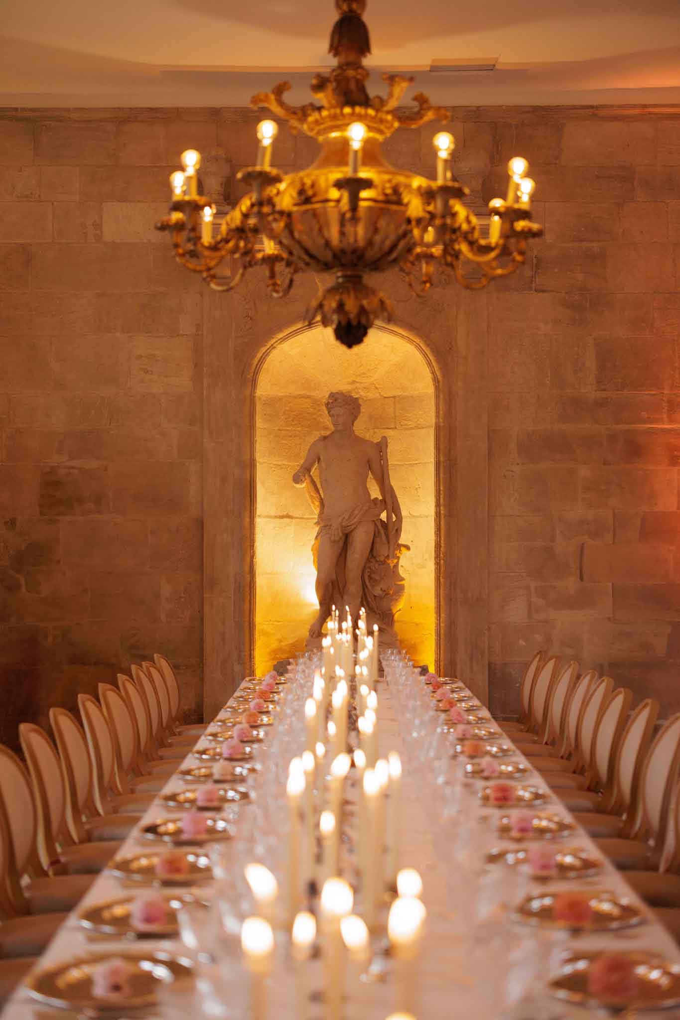 A reception dinner table setup photographed from one end looking down the full length of the table toward an arched stone niche housing a classical figurative sculpture, lit with warm amber uplighting. The long rectangular table is set for approximately 30 guests, lined on both sides with upholstered cream dining chairs. The table runner features densely packed ivory taper candles and small votive candles interspersed with scattered pink floral blooms, creating a continuous line of candlelight down the center. Gold charger plates and glassware are visible at each place setting. A large ornate gilt candelabra-style chandelier hangs in the foreground. The room features exposed limestone ashlar walls and a classic French chateau interior style. Wide shot taken from a low central angle emphasizing the table's length and the sculptural focal point at the far end. Potential venue feature image.