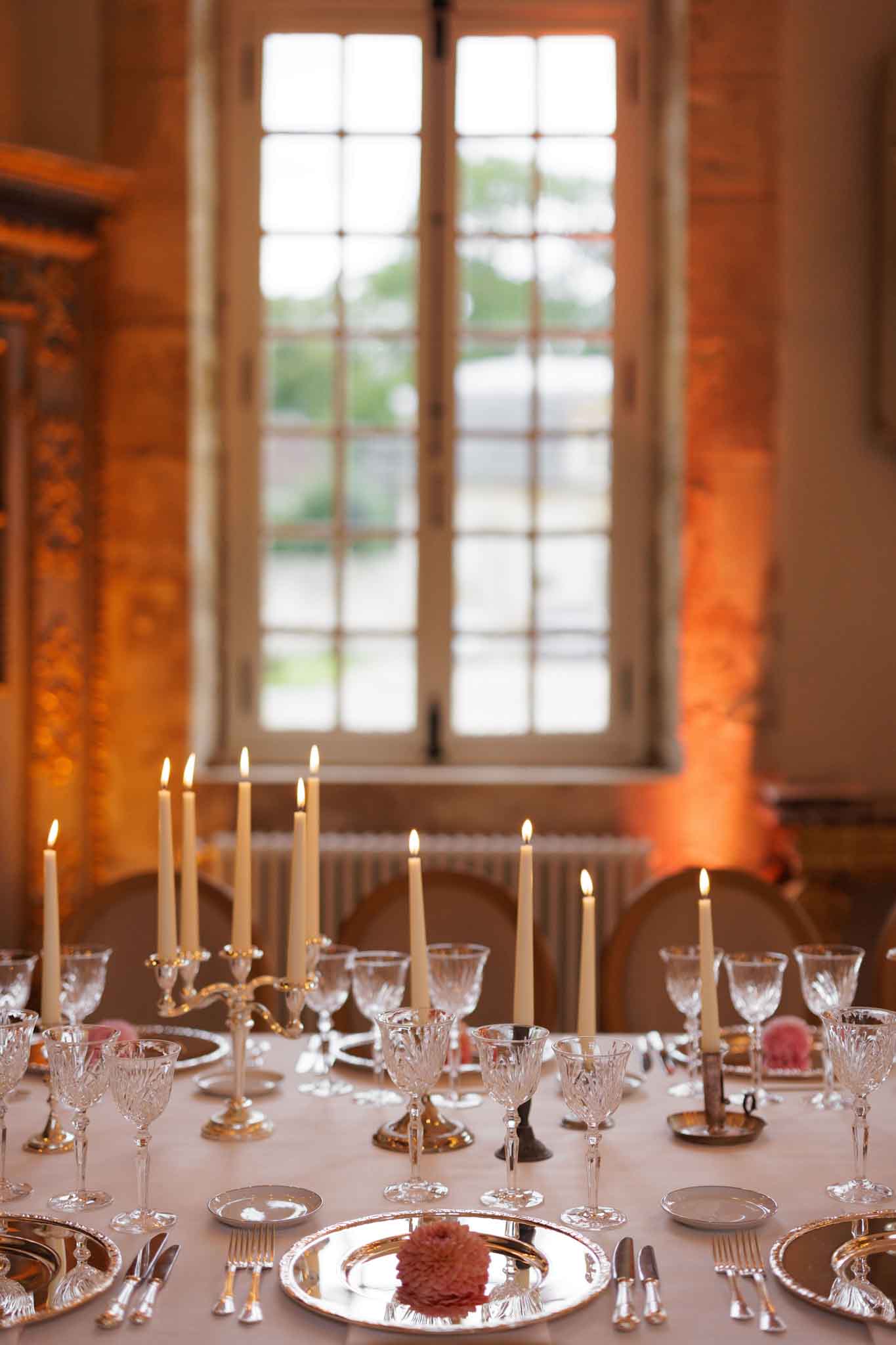 A close-up detail shot of a wedding reception table set inside what appears to be a chateau or historic manor room, featuring tall grid-paned windows and ornate gilded wall paneling visible in the background. The table is dressed with a cream linen cloth and set with silver charger plates, crystal cut-glass stemware in multiple sizes, and silver flatware. Ivory taper candles in silver candelabras and individual silver candlestick holders are lit across the table, casting warm amber light that is complemented by orange uplighting on the walls. Small dusty pink flower blooms — likely carnations or dahlias — are placed on the silver charger plates as individual napkin or place decorations. The overall decor palette is ivory, silver, and blush pink with a classic, formal French style.