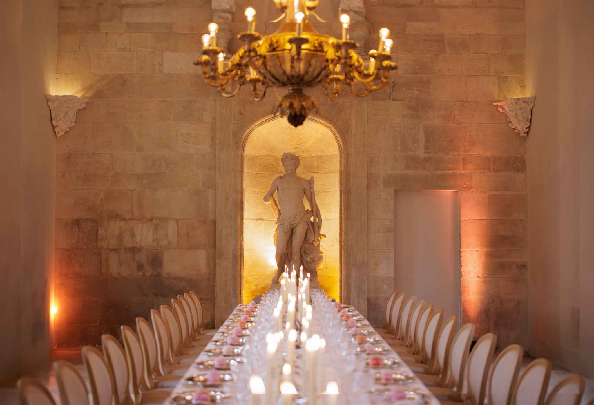 A wedding reception dinner setup photographed from one end of a long single banquet table stretching toward the far wall of an indoor stone hall. The table is lined on both sides with white French Louis-style chairs and set with glassware, gold-toned place settings, and small pink floral accents at each place setting. A continuous row of lit taper candles runs the full length of the table's center. The room is lit with warm amber uplighting along the stone block walls and a large gold chandelier with exposed bulbs hanging overhead. At the far end, a classical marble-style figurative statue is displayed in a illuminated arched stone alcove, serving as a focal point down the table. The decor palette combines warm gold, white, and blush pink in a classic French chateau style. Wide shot taken from table level looking straight down the length of the room. Potential venue feature image.