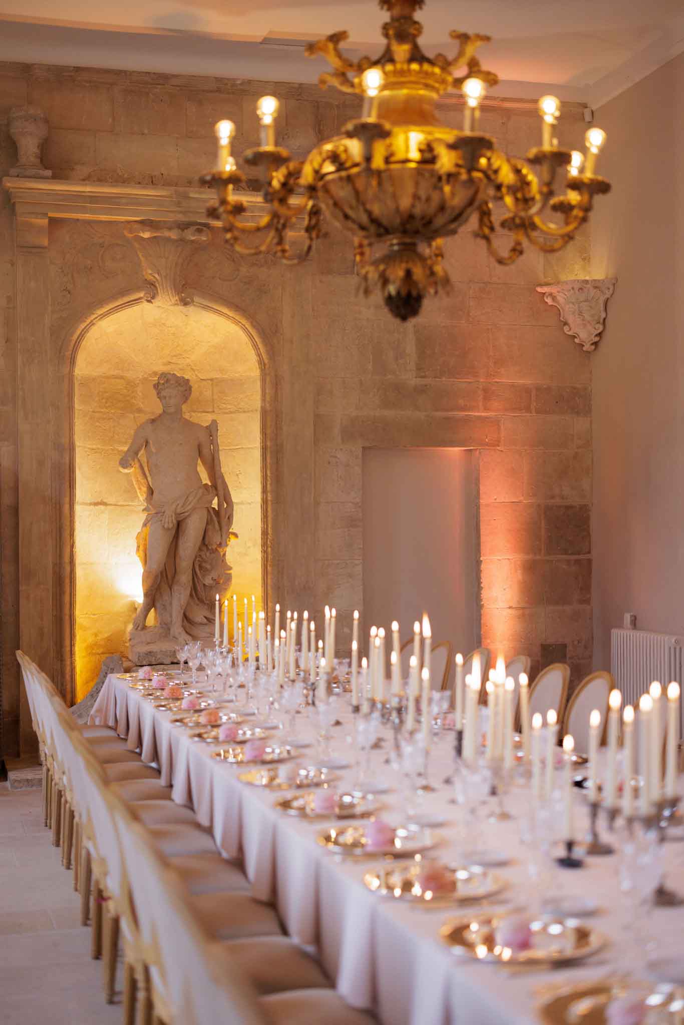 A wedding reception dinner table setup photographed indoors in what appears to be a French chateau ballroom or grand hall, with no guests present. A long rectangular table is dressed in white linen and set with gold charger plates, crystal glassware, and scattered small pink floral or decorative elements at each place setting. Dozens of tall ivory taper candles on individual candlesticks run the length of the table, casting warm ambient light. Gold-framed upholstered chairs line both sides of the table. The room features exposed limestone walls, a classical stone niche housing a full-length figurative sculpture lit from below with warm uplighting, and a large ornate gold chandelier with exposed Edison-style bulbs hanging overhead. Additional warm pink-toned uplighting illuminates the far wall. The composition is a wide angled shot taken from one end of the table looking toward the sculpture niche, with shallow depth of field. Potential venue feature image.