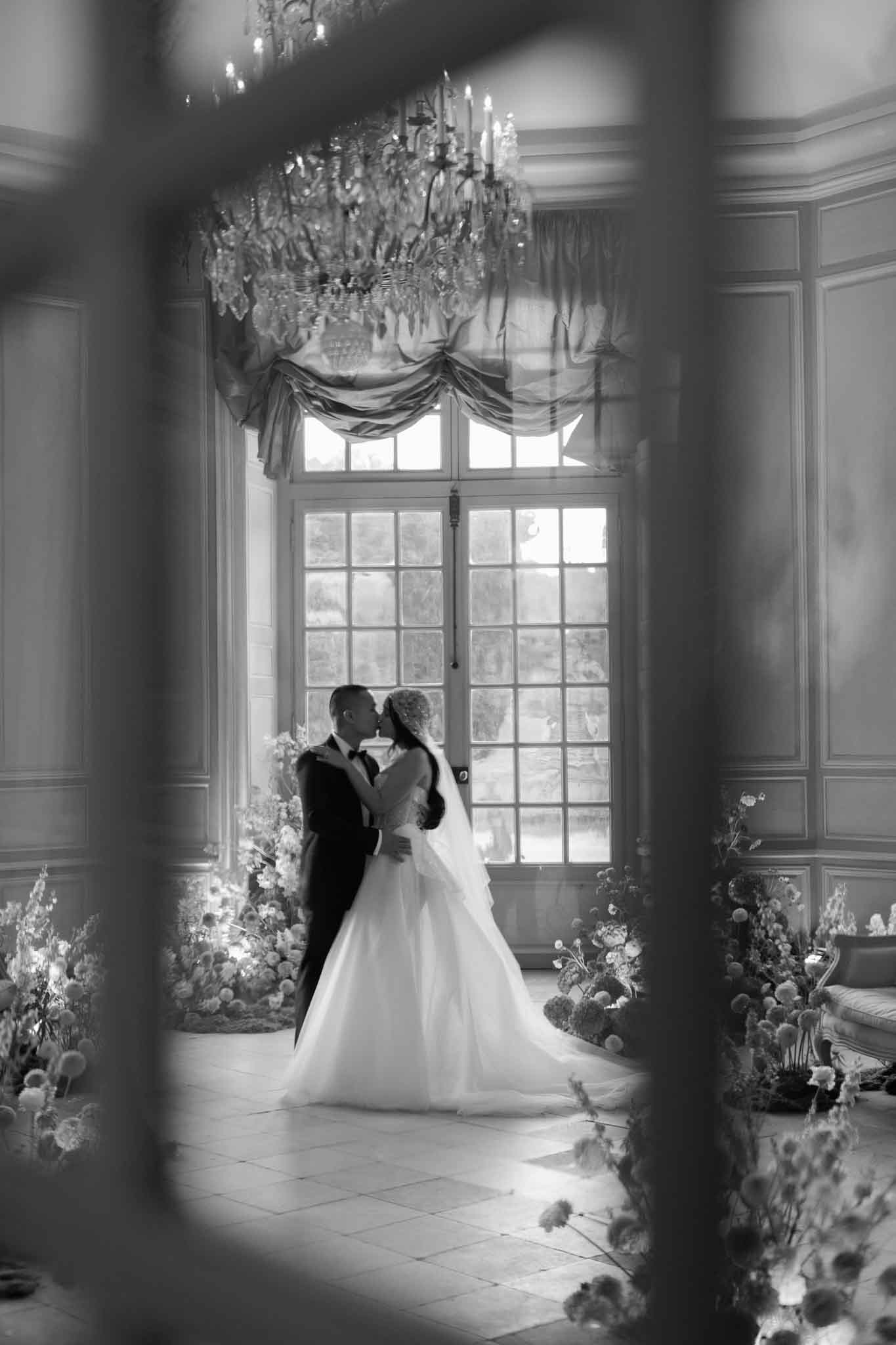Black-and-white portrait of a bride and groom sharing a kiss inside a formal chateau salon, shot through a doorway or architectural frame that creates a natural vignette around the edges. The groom wears a dark tuxedo with bow tie, and the bride wears a full ballgown with a long train and what appears to be a floral hair accessory. They are positioned in front of tall French double doors with grid-pane glazing and draped swag curtains, with natural light backlighting the couple. A large crystal chandelier with candle-style arms hangs prominently above, and the room features classic wood-paneled boiserie walls and a tiled floor. Large loose floral arrangements — appearing to include globe thistles, dahlias, and airy wildflower-style blooms — are placed in clusters on the floor flanking the couple on both sides, with warm spotlighting highlighting the florals against the darker room. The composition is a medium-wide portrait framed through an architectural opening, with high contrast tones emphasizing the backlit window and the bright gown against the darker surroundings. Potential venue feature image.