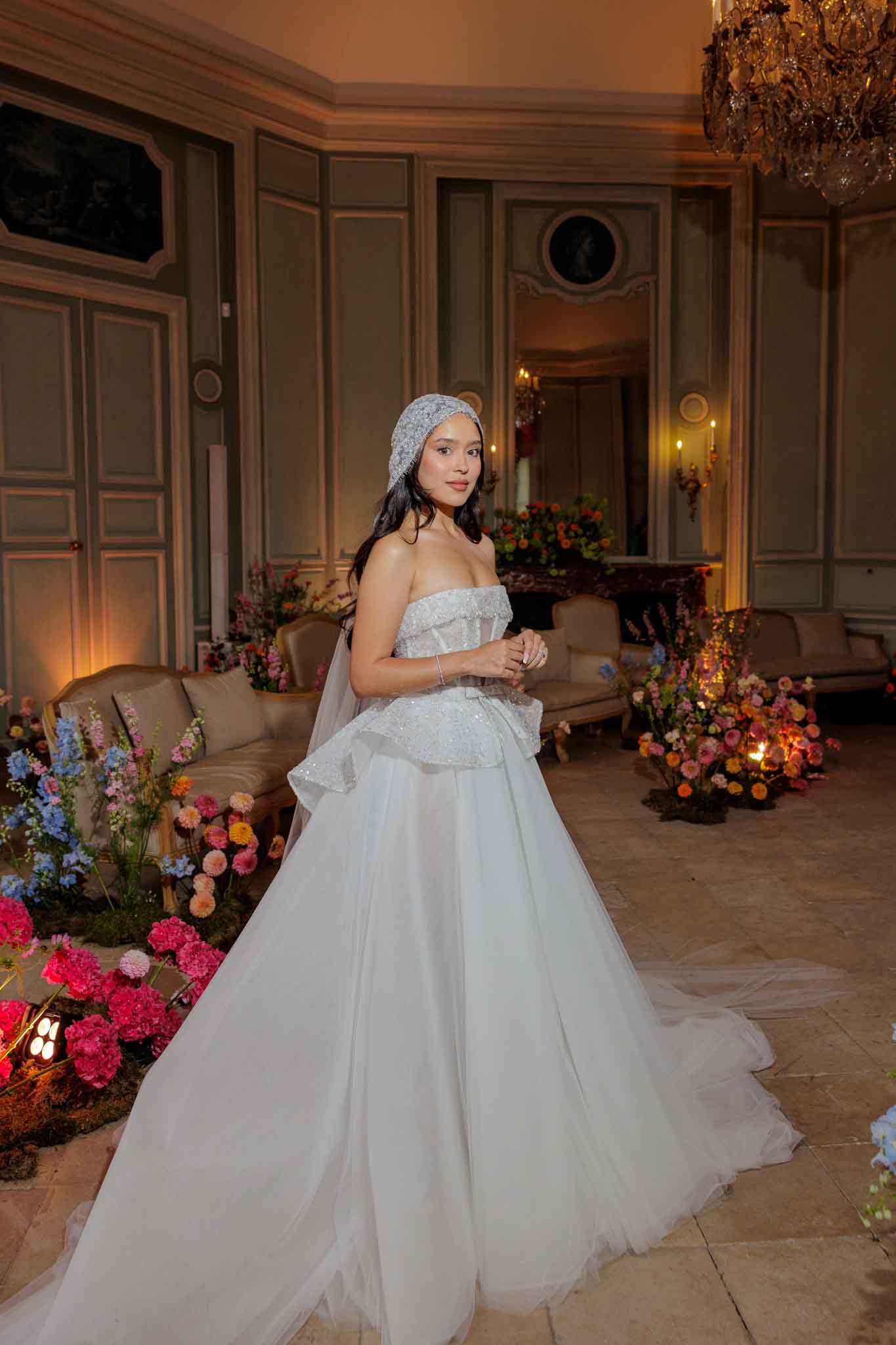 A bride stands alone in a portrait shot inside what appears to be a French chateau salon or reception room, lit by warm ambient lighting and a crystal chandelier overhead. She wears a strapless white ballgown with a sequined peplum bodice and a full tulle skirt with a train, paired with a beaded or sequined headband-style bridal cap and a sheer veil. The room is decorated with abundant low garden-style floral arrangements in vibrant mixed colors including hot pink, coral, blue, orange, and burgundy flowers placed directly on the floor around the perimeter, alongside candles and uplighting. The paneled walls are painted in a muted sage green with ornate classical molding, and period-style upholstered seating is visible in the background.