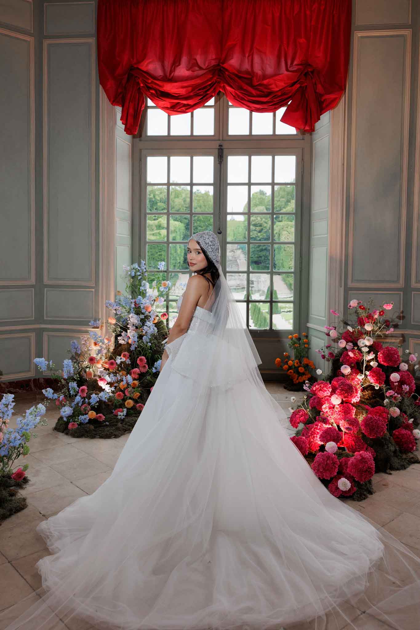 A bride poses indoors in what appears to be a French chateau ballroom or grand salon, standing near a large multi-pane window draped with a deep red silk swag valance. She wears a strapless white tulle ballgown with a voluminous cathedral-length train, paired with a lace-edged bridal bonnet-style headpiece and a sheer veil. She is turned away from the camera, glancing back over her shoulder. Flanking her on both sides are large floor-level floral installations arranged on beds of moss: the left cluster features periwinkle blue delphiniums, coral and peach ranunculus, and orange blooms, while the right cluster is composed of deep crimson and hot pink peonies, carnations, and white anemones, lit with warm accent lighting embedded in the arrangements. The room features pale grey-green painted paneling. Through the window, a formal French garden with manicured hedgerows is visible in the background. Full-length portrait shot.