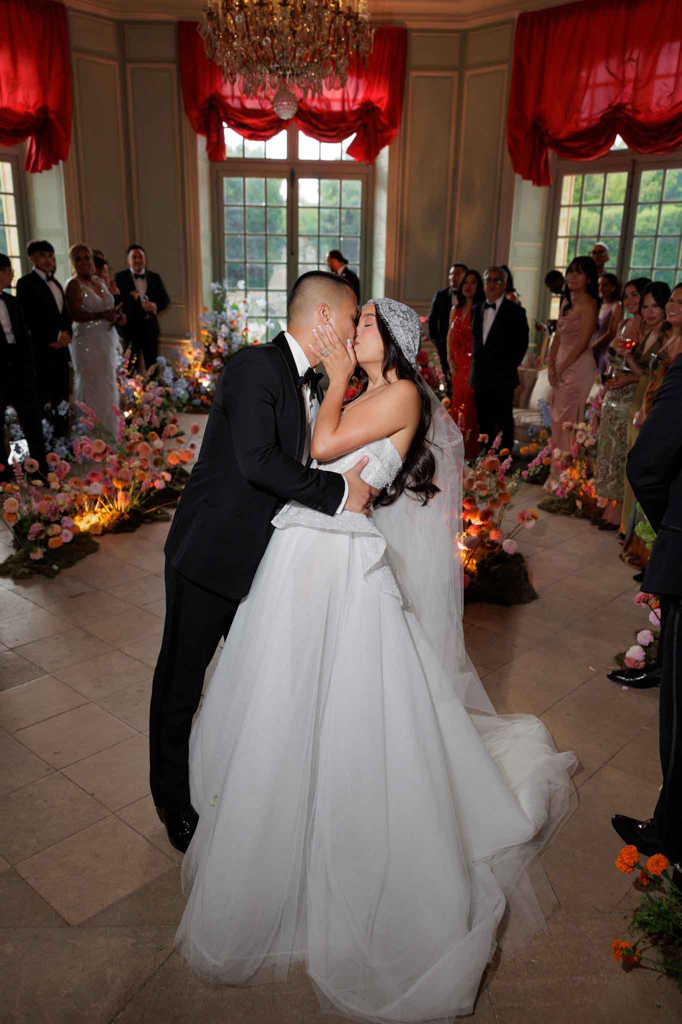 The bride and groom share their first kiss during an indoor wedding ceremony inside what appears to be a French chateau salon. The groom wears a black tuxedo with a bow tie, and the bride wears a white strapless ball gown with a voluminous tulle skirt, a beaded off-shoulder detail, and a lace-edged veil with an ornate beaded headpiece. The room features tall French windows with dramatic red silk swag drapery and a crystal chandelier overhead, with stone tile flooring. Lining the aisle on both sides are lush ground-level floral arrangements in a warm, jewel-toned palette of coral, orange, pink, peach, and lavender blooms including ranunculus, dahlias, and garden roses, warmly lit from below. Approximately 20–30 guests in formal attire — including black tuxedos and colorful gowns in red, pink, and gold — stand watching on either side. The shot is a full-length portrait taken from a low angle, capturing the full sweep of the bride's dress and train.