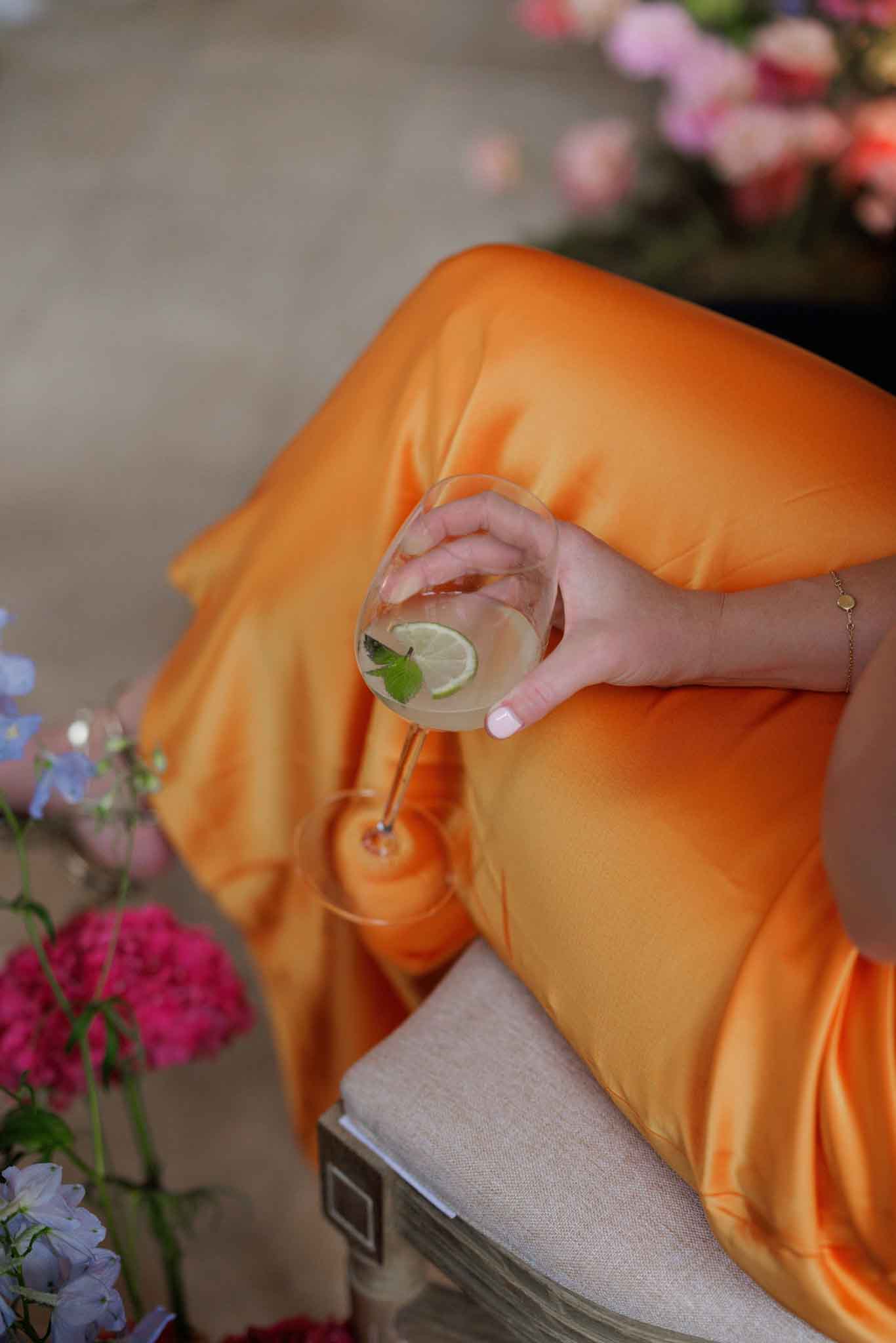 Close-up detail shot of a guest or bridesmaid during what appears to be a cocktail hour or reception, seated on a linen-upholstered chair. The subject is wearing a vivid orange satin dress and holds a stemmed gin balloon glass containing a clear cocktail garnished with a lime slice and fresh mint. A delicate gold chain bracelet is visible on her wrist. Surrounding the chair are loose florals including hot pink peonies or stock flowers and soft blue delphinium, with pink roses visible in soft focus in the background, suggesting a colorful, garden-inspired floral palette.