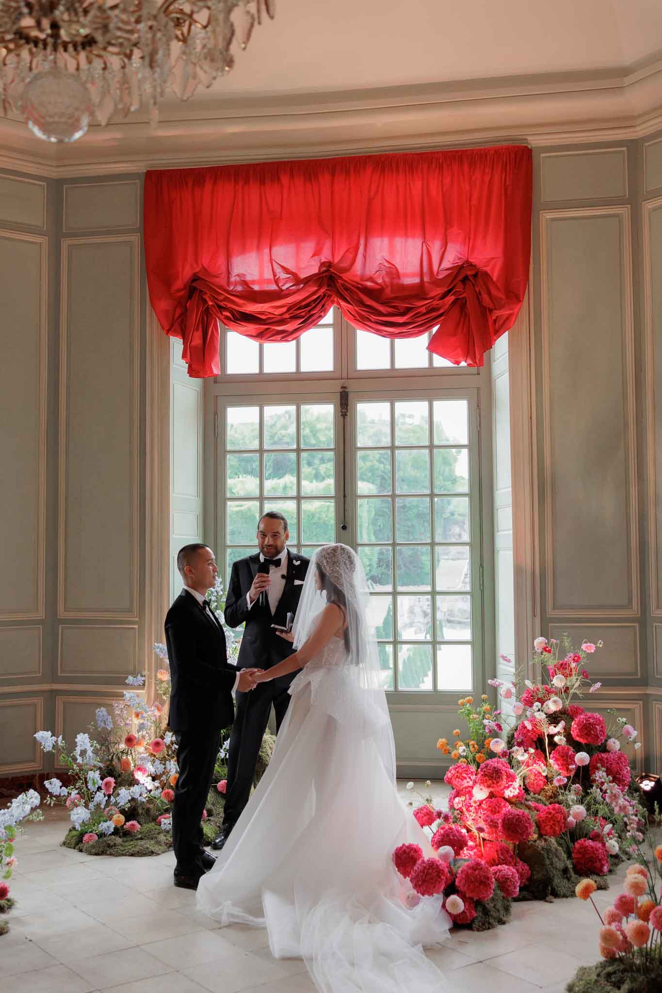An indoor wedding ceremony taking place in a classical French chateau room with sage-green paneled walls, ornate crown molding, and a crystal chandelier overhead. The couple stands facing each other holding hands before an officiant dressed in a black tuxedo holding a microphone, all positioned in front of a tall multi-pane window dressed with a draped red silk balloon valance. The bride wears a white ballgown with a cathedral-length veil featuring lace detailing at the crown, while the groom wears a black tuxedo. The ceremony altar area is framed by two large ground-level floral installations: one on the right composed densely of hot pink and magenta dahlias, blush pompons, peach ranunculus, and white blooms, and one on the left featuring loose arrangements of lavender-blue delphinium, peach, and coral wildflower-style stems with moss, creating a bold asymmetrical color contrast. Wide-shot portrait composition capturing all three figures and the full ceremonial setting.