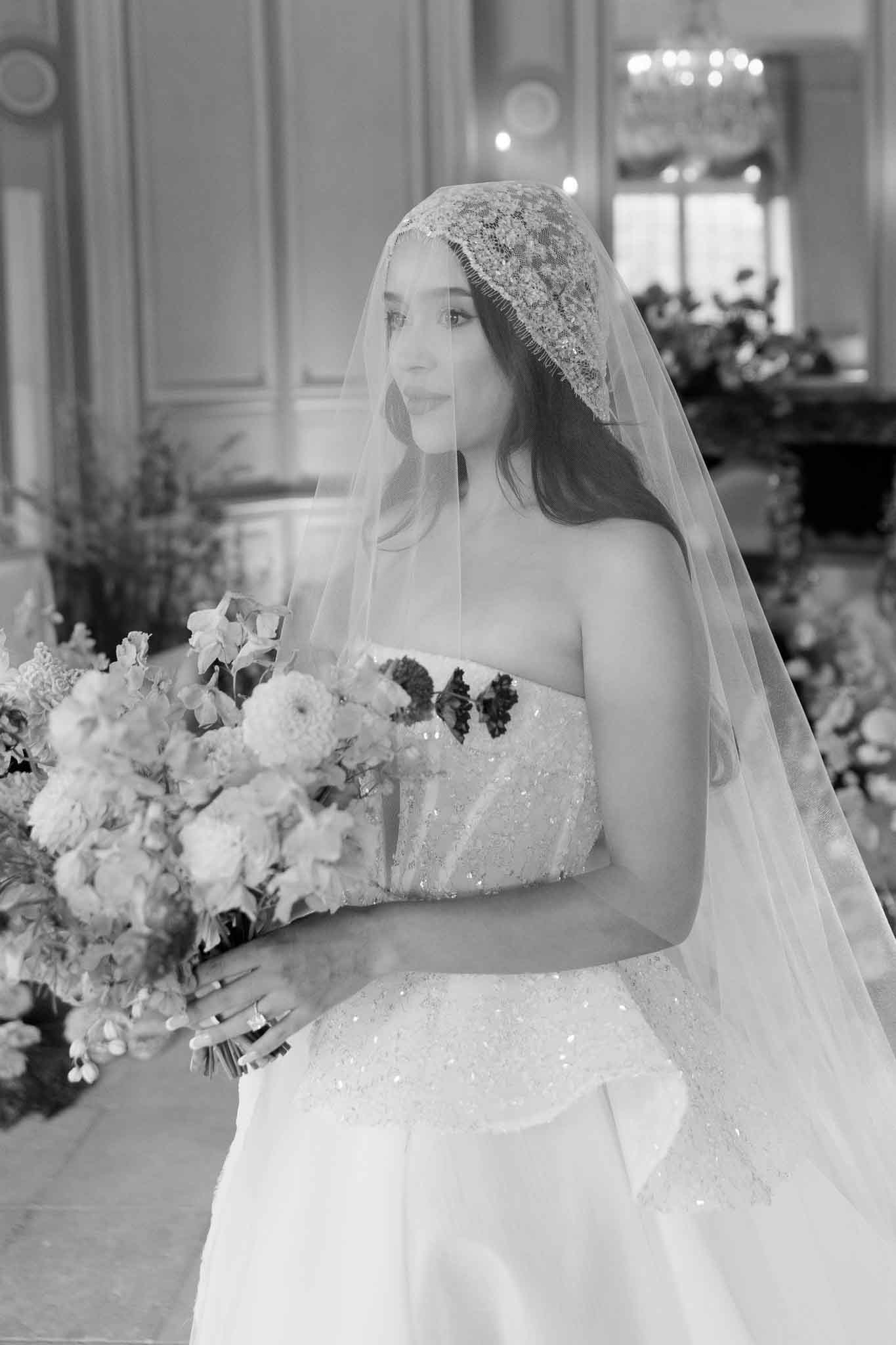 A black-and-white bridal portrait of a bride standing indoors in what appears to be a classic French chateau or mansion interior, identifiable by ornate wood-paneled walls and a chandelier visible in the background. The bride wears a strapless ball gown with heavily beaded and sequined bodice detailing, paired with a Juliet-cap veil featuring intricate lace appliqué and a delicate beaded edge. She holds a large, lush bouquet featuring what appear to be dahlias, ranunculus, sweet peas, and darker accent blooms, rendered in soft light and dark tones in the monochrome image. The composition is a mid-length portrait shot slightly from the side, with the bride gazing off-camera; large floral arrangements are softly visible in the background, consistent with a classic, formal wedding aesthetic.