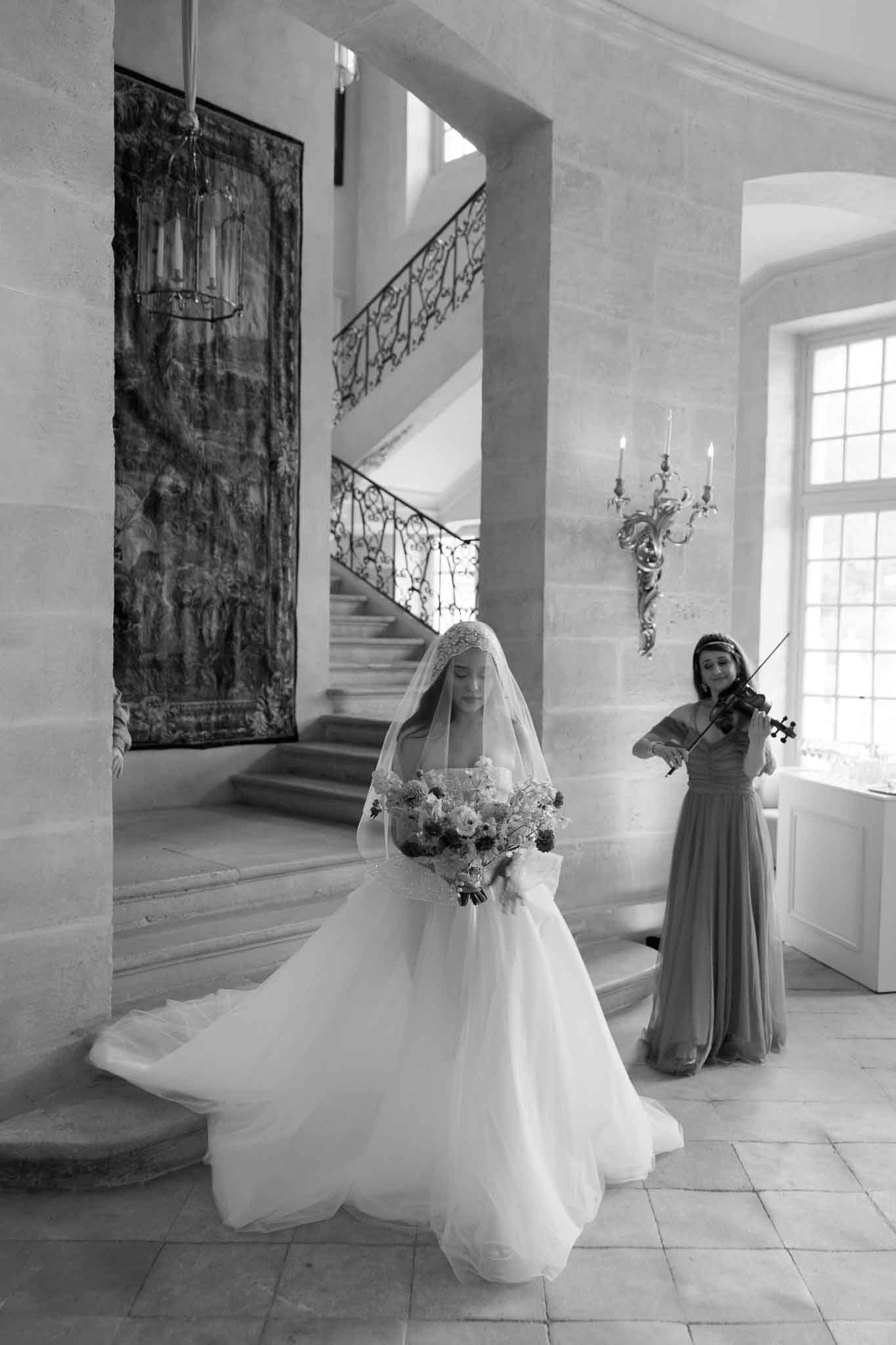 Black-and-white portrait of a bride standing in the grand interior entrance hall of a French chateau, holding a full rounded bouquet of mixed blooms and looking downward with eyes closed. She wears a full-skirted ballgown with a long cathedral-length veil featuring a lace edge, and the gown has an extended train spread across the stone tile floor. In the background to the right, a female musician in a floor-length dress plays violin near a tall window. The space features a sweeping wrought-iron staircase, a large decorative tapestry hung on the stone wall, a hanging candle lantern, and an ornate gilded wall sconce with lit candles. The image has bright highlights near the window and deep shadows in the staircase area, giving it strong tonal contrast. Potential venue feature image.